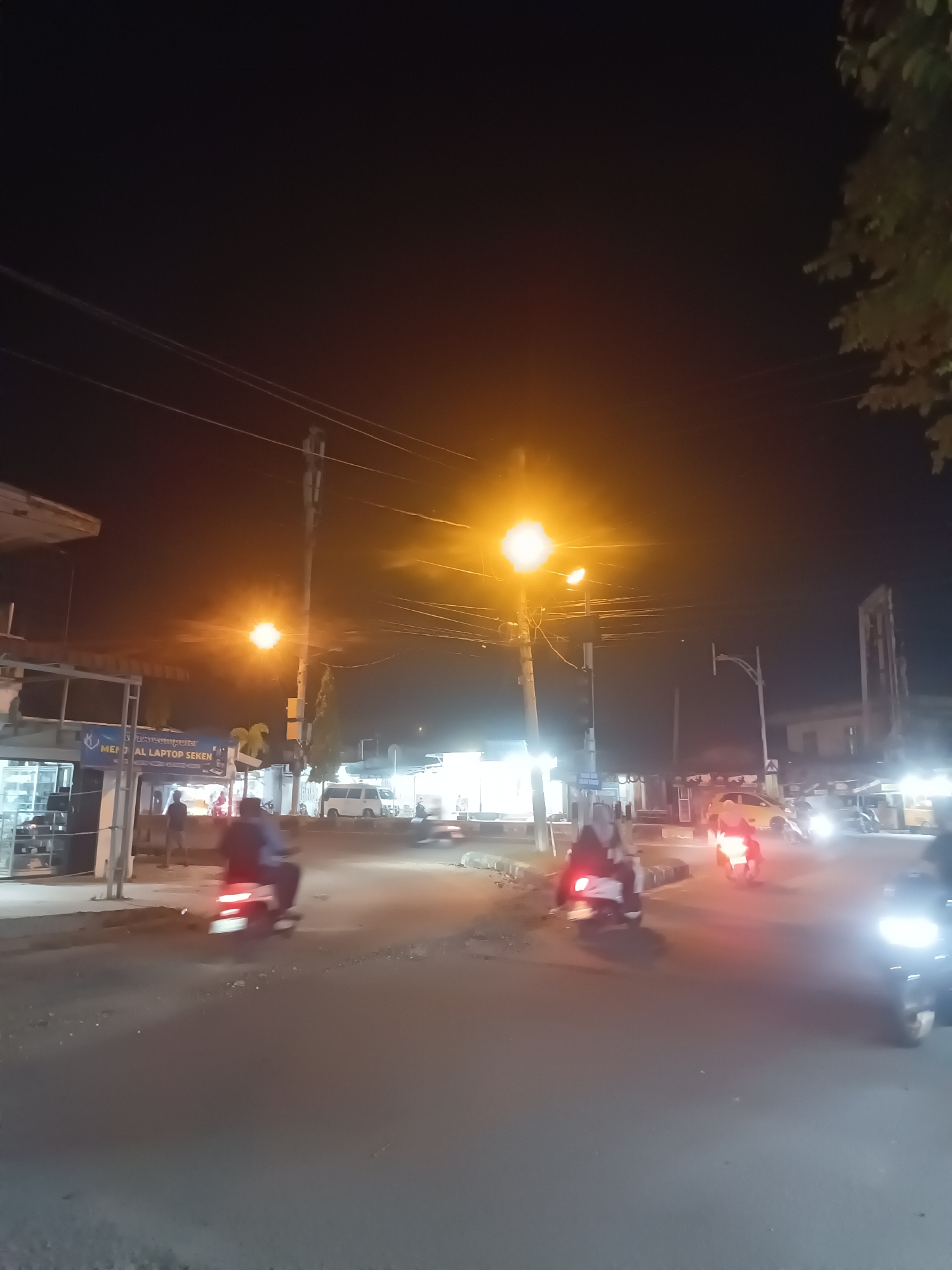 This is a nighttime street scene in Lhokseumawe, Indonesia. The image captures a road with several motorbikes, some of which are in motion, their headlights and taillights creating streaks of light. There are streetlights illuminating the scene with a warm glow. On the left side of the road, a building with storefronts is visible, one displaying a sign that appears to say "MEMORIAL LAPTOP SEKEN." Pedestrians can be seen in front of the storefronts. The overall environment suggests a commercial or residential area with active street life at night.