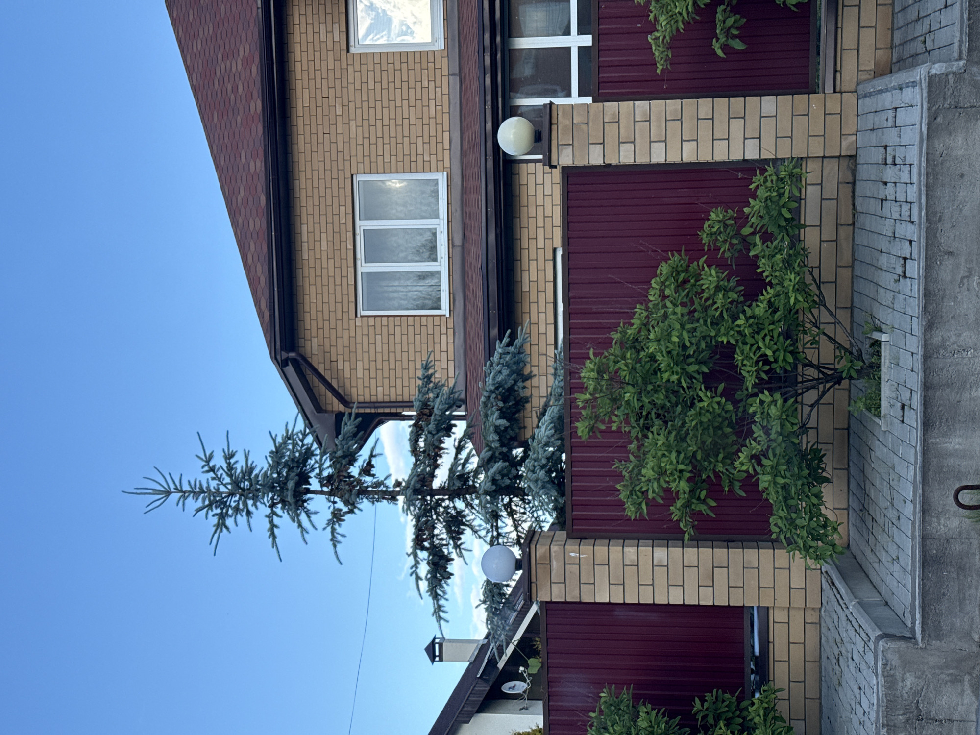 In the city of Prikamskiy, Russia, a two-story brick house with a maroon shingle roof is pictured. The house has two visible windows on the upper floor, both with white frames and curtains. A tall, slender evergreen tree with blue-green needles stands in front of the house, partially obscuring the upper portion of the structure.
A dark red metal fence with light tan brick pillars separates the property from the foreground. Each pillar is topped with a white, spherical lamp. In front of the fence, a small, leafy green tree is planted in a paved area. The paving stones are arranged in a geometric pattern. The foreground consists of a concrete structure with a metal ring embedded in it. The sky is a clear blue with a few wispy clouds. The lighting suggests it is late afternoon or early evening.