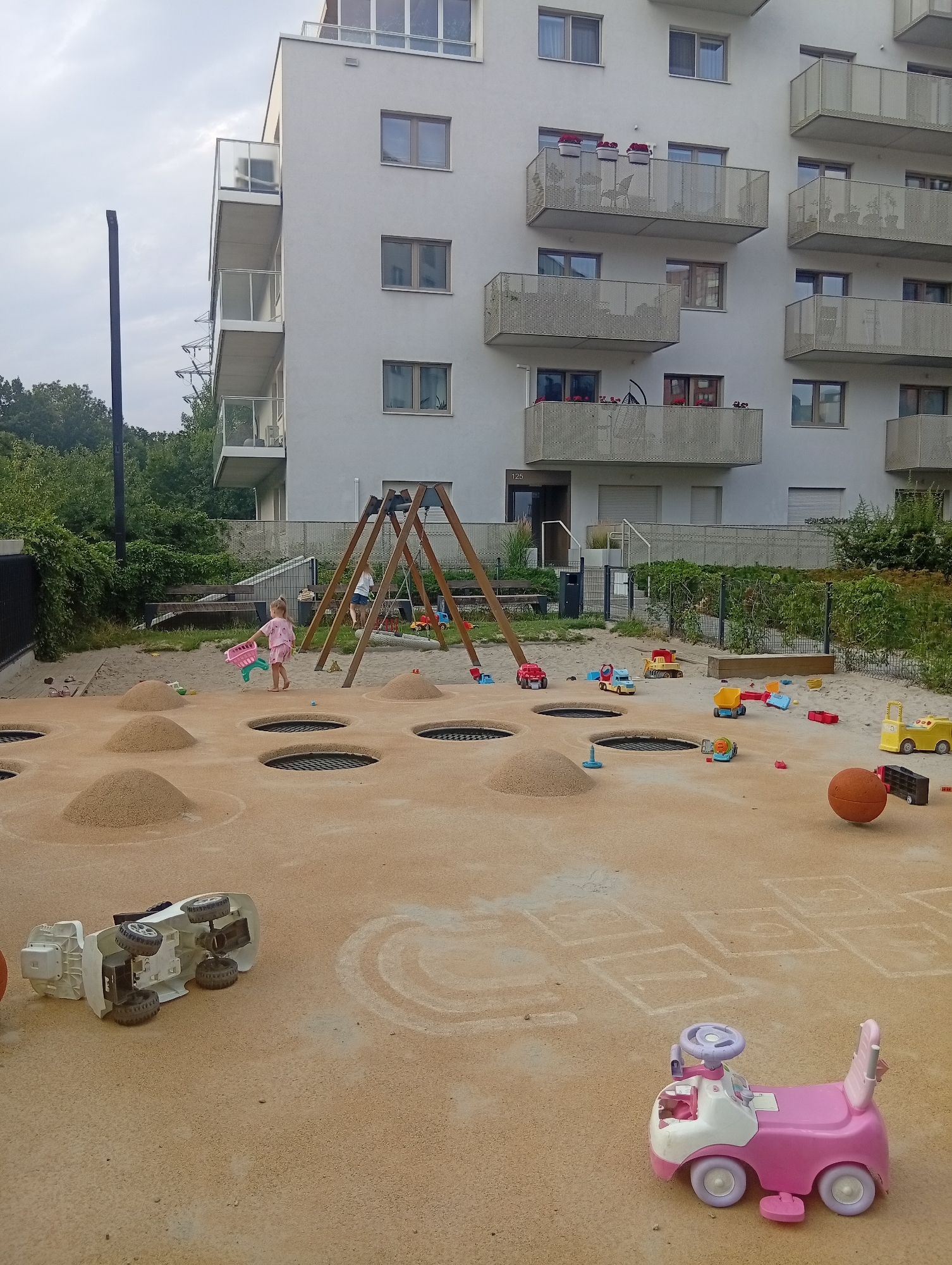 The image depicts an outdoor playground area adjacent to a modern apartment building. The playground is covered in sand and features several circular, sunken trampolines. Scattered across the sand are various children's toys, including small trucks, a partially overturned white toy car, a pink and white ride-on toy car, and a collection of building blocks. In the background, a wooden swing set with two swings is visible, and a young girl in pink clothing is holding a pink toy in her hand. Another child, obscured by the swing set, is also present. The apartment building is a multi-story structure with balconies and windows. Trees and foliage surround the playground area, and a metal fence runs along the perimeter. The sky is overcast. Based on the context of this scene, it is highly likely located in Wrocław, Poland, a city known for its contemporary residential developments and green urban spaces.