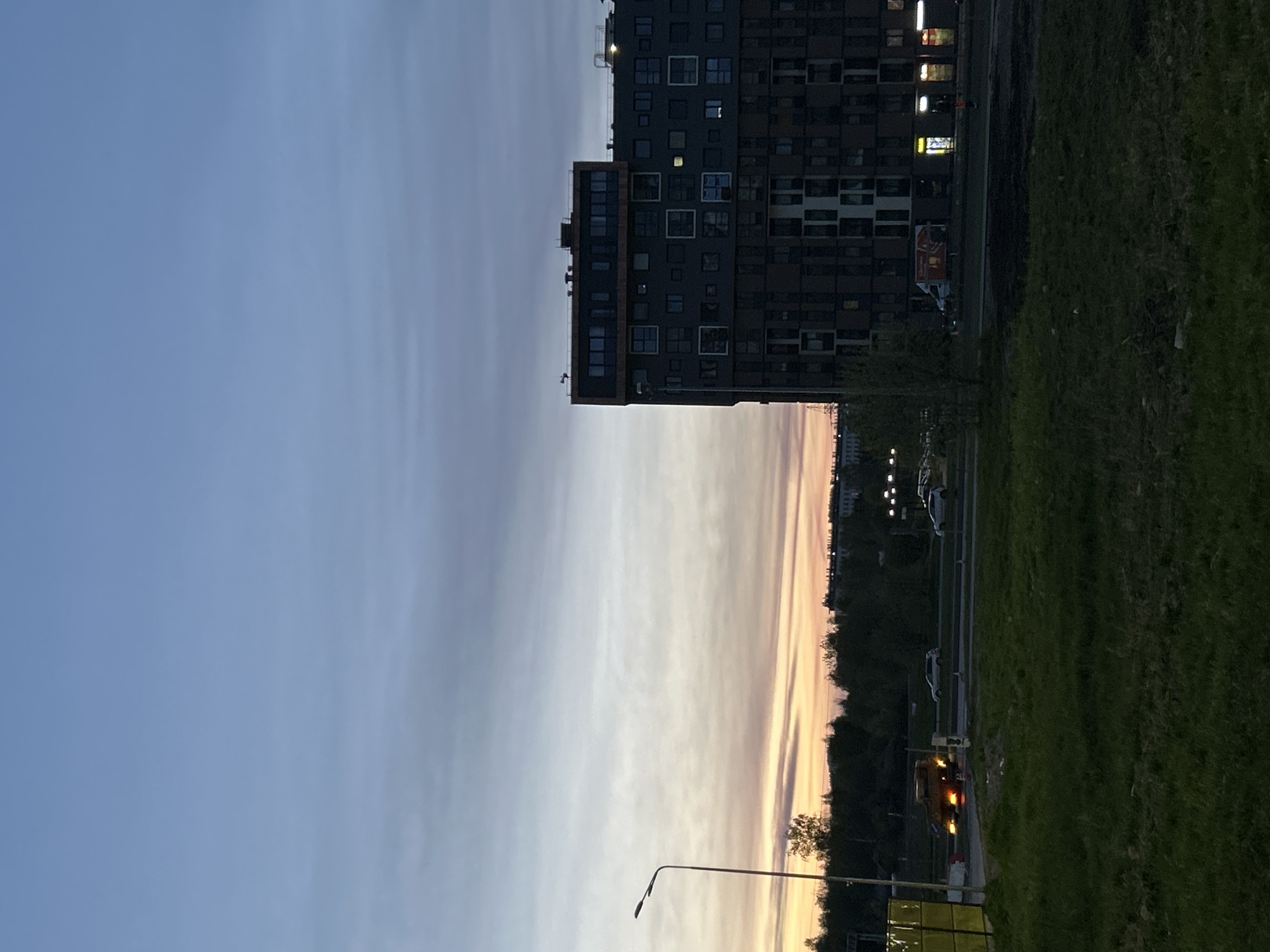 The image shows a view in Saint Petersburg, Russia, at dusk or dawn. The sky is transitioning from blue to shades of pink and orange near the horizon. A modern high-rise building is visible on the right, with multiple windows and lights on some floors. A street with a row of trees separates the building from a grassy area. Cars are parked along the street. A street lamp is on the lower left.