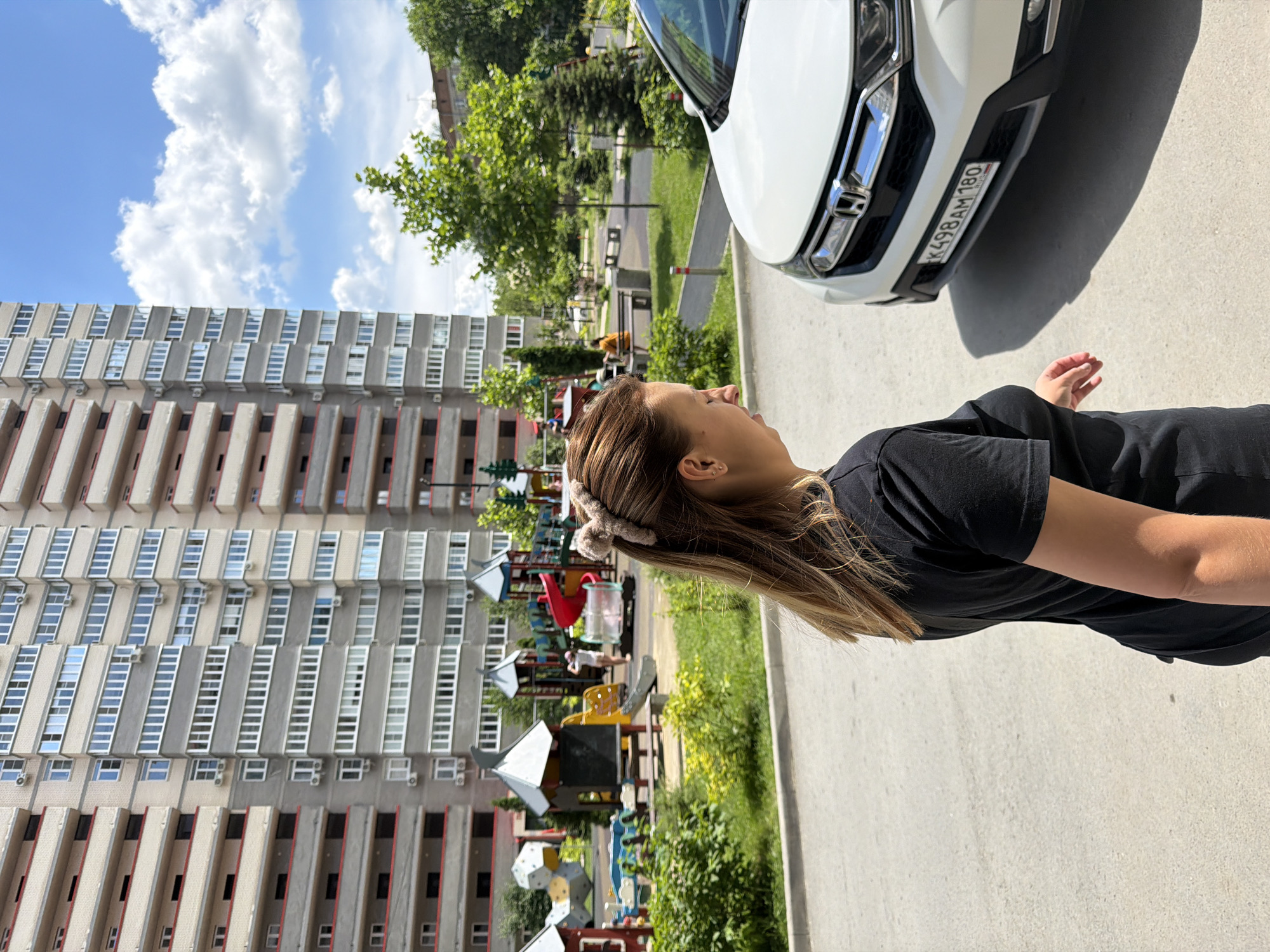 A woman with brown hair, wearing a black t-shirt, stands in the foreground of the image, partially obscuring a white car and a playground. She is facing to the right, with her head tilted down. The car, a Honda CR-V, is parked on an asphalt surface. Behind the woman and car, a playground with various structures, including slides and climbing equipment, is visible. In the background, a tall, multi-story apartment building dominates the scene. The sky above is blue with scattered white clouds. The setting appears to be a residential area within the city of Ovoshchnoy, Russia.