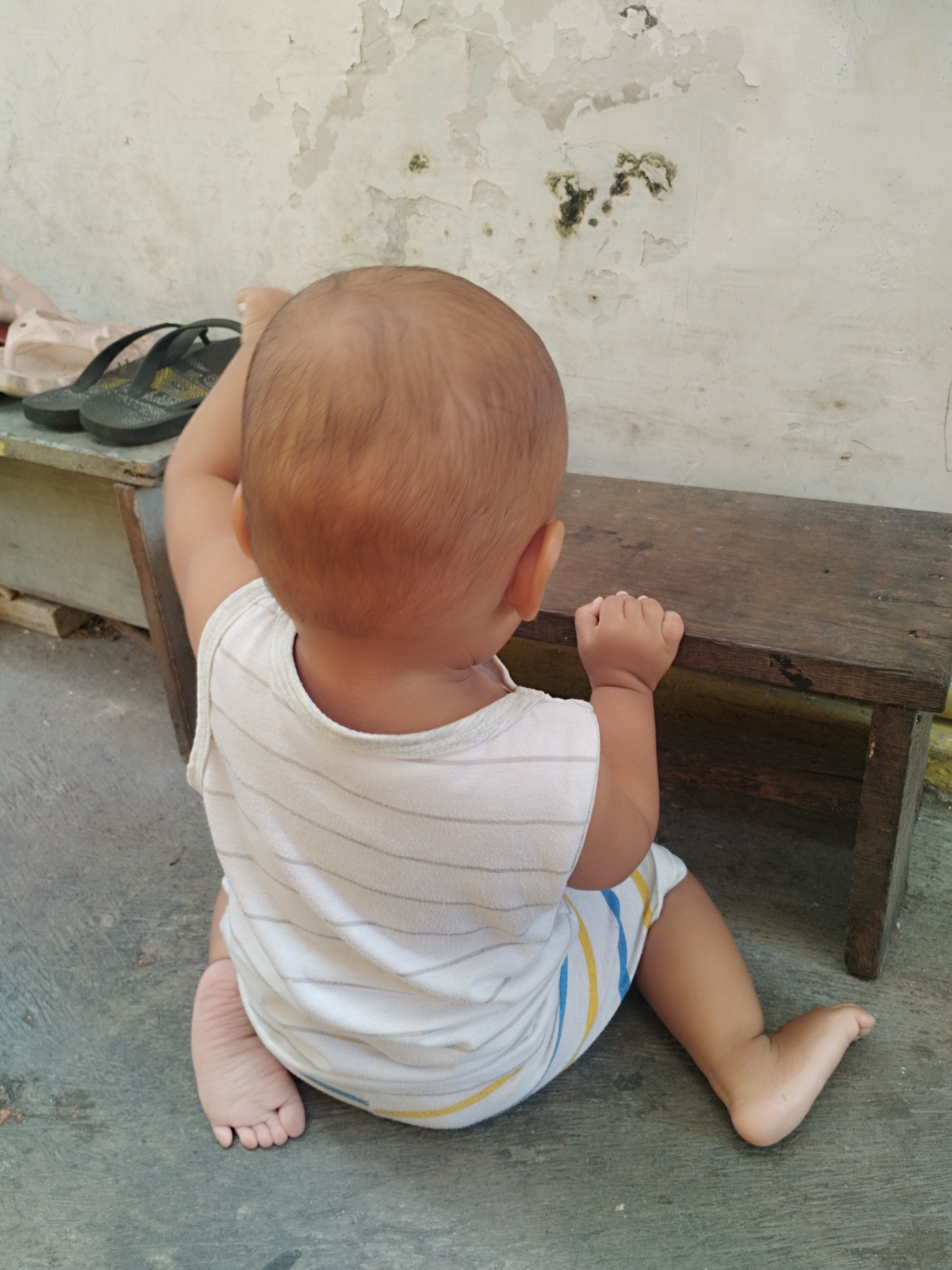 A toddler sits on a concrete floor in front of a wooden bench, facing away from the camera. The toddler wears a white tank top with thin horizontal stripes and shorts with vertical blue and yellow stripes. The toddler's left leg is bent and positioned behind them, while their right leg is extended forward. Their left arm is raised, and their hand is placed on the back of their head, while their right hand rests on the bench. The background shows a weathered, off-white wall with peeling paint and green algae patches. To the left of the toddler, on a shelf, are a pair of pink sandals and a pair of black flip-flops. The scene appears to be an outdoor area, possibly a yard or alleyway, located in Surabaya, Indonesia, suggested by the architecture and climate inferred from the environment.