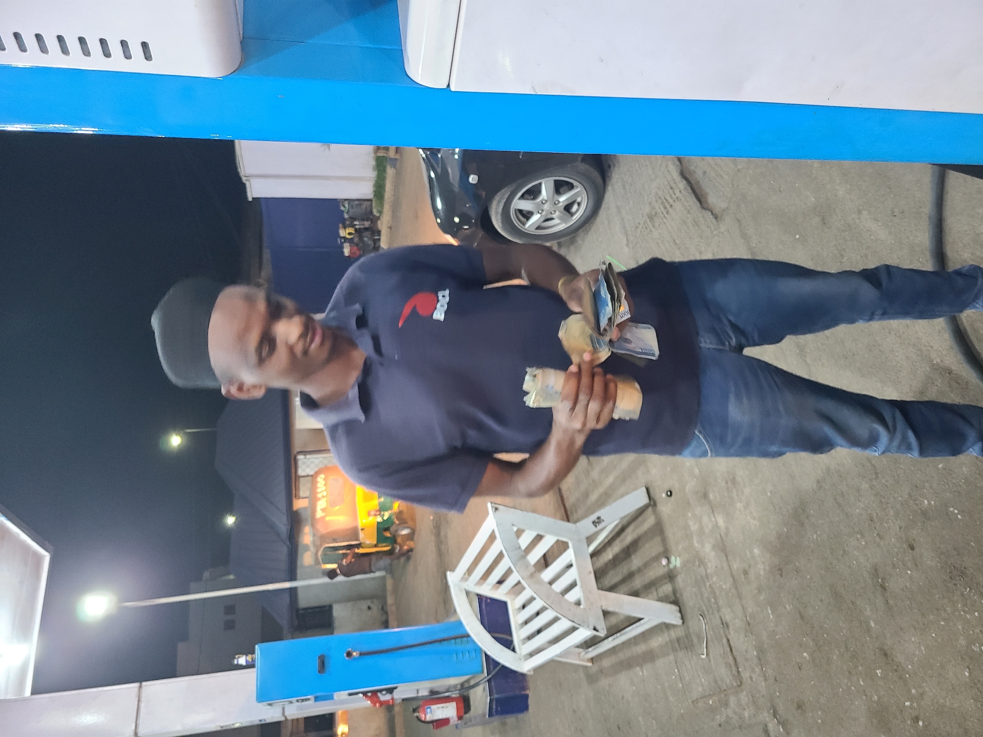 The image shows a man holding cash in front of a petrol station in Jos, Nigeria at night. The man wears a dark blue polo shirt, jeans, and a black cap. He is standing on a concrete surface beside a turned-over white chair. In the background are other petrol pumps, a parked black car, and lit-up buildings including one with the sign "PETROL #1600". Street lights illuminate the area.