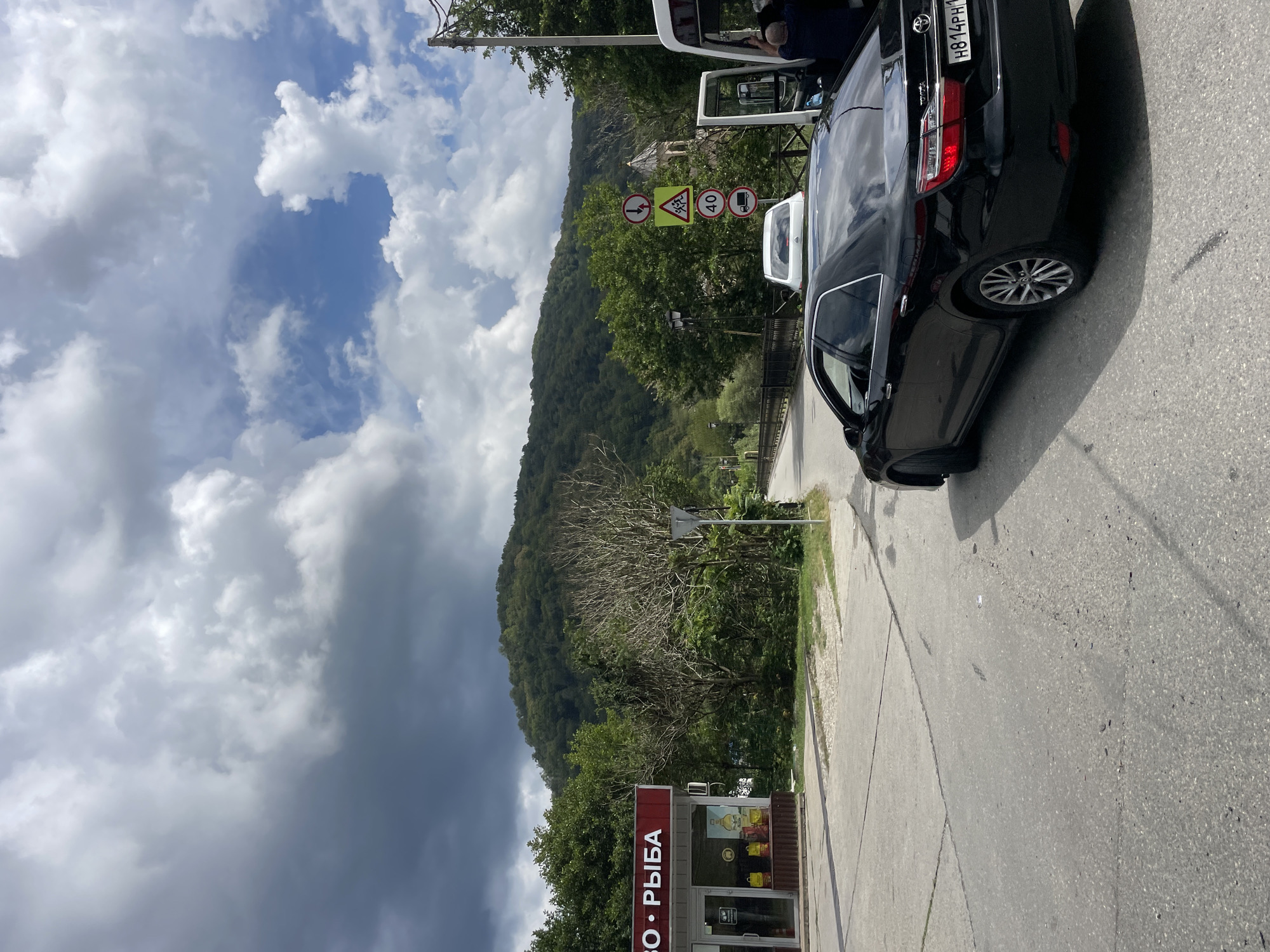 A black sedan is parked on an asphalt road. A white van is parked next to it, with its doors open and a person visible inside. A building with a red awning, possibly a shop, is on the left side of the road. In the background, a lush green mountain rises under a cloudy sky. Traffic signs are visible near the van, indicating a speed limit of 40 and a pedestrian crossing. The scene appears to be in Plastunka, Russia, based on the context provided.