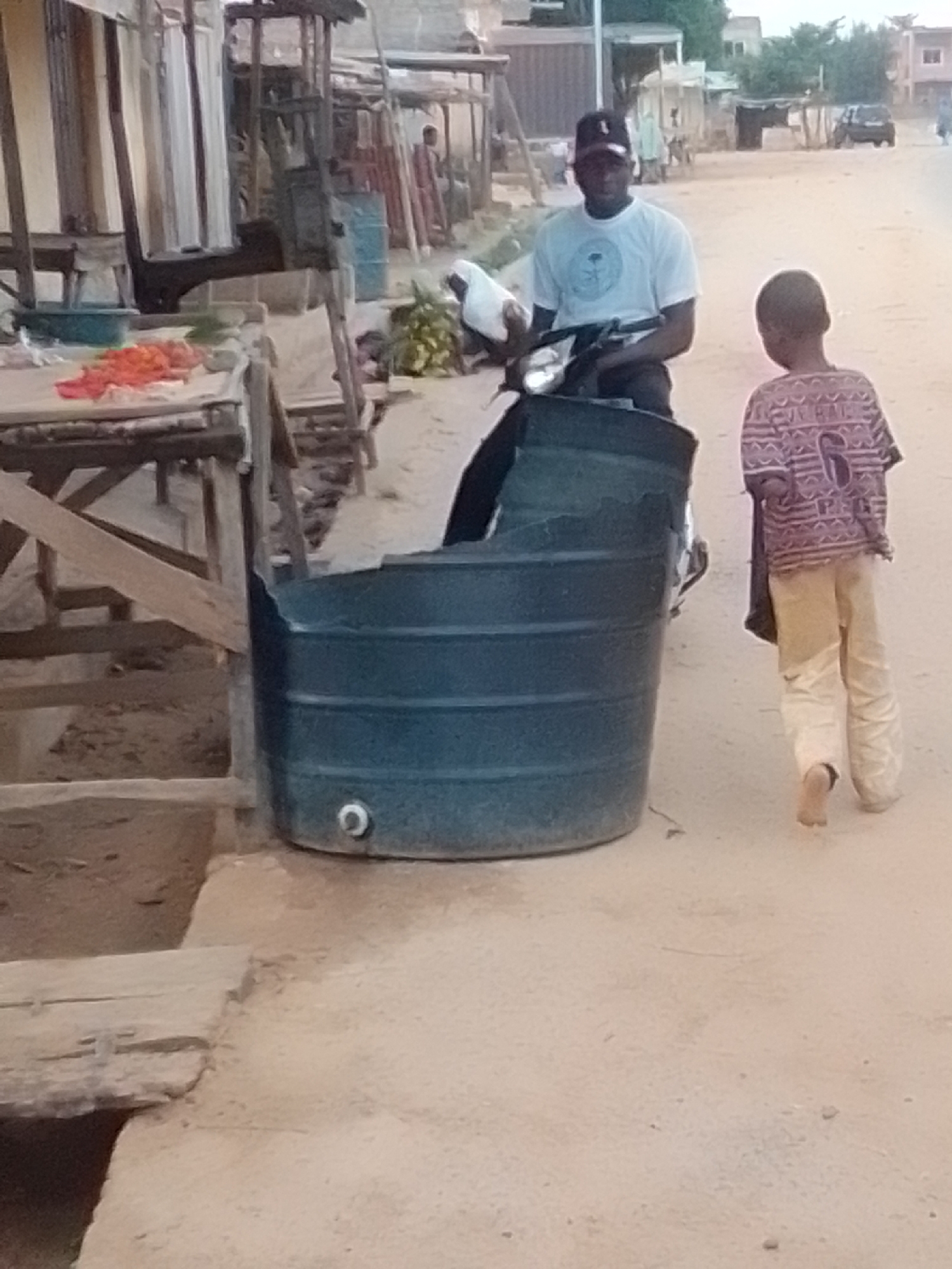 In Galadima, Nigeria, a man wearing a white t-shirt and a black baseball cap is seated on a motorbike. Beside him, a child with short dark hair, wearing a patterned long-sleeved shirt and light-colored pants, walks away from the camera. In the foreground, a large, dark blue plastic water tank is positioned on the ground. The environment appears to be an outdoor market setting, with wooden stalls and various goods, including red produce, visible in the background. The ground is covered in a light-colored, dusty earth.