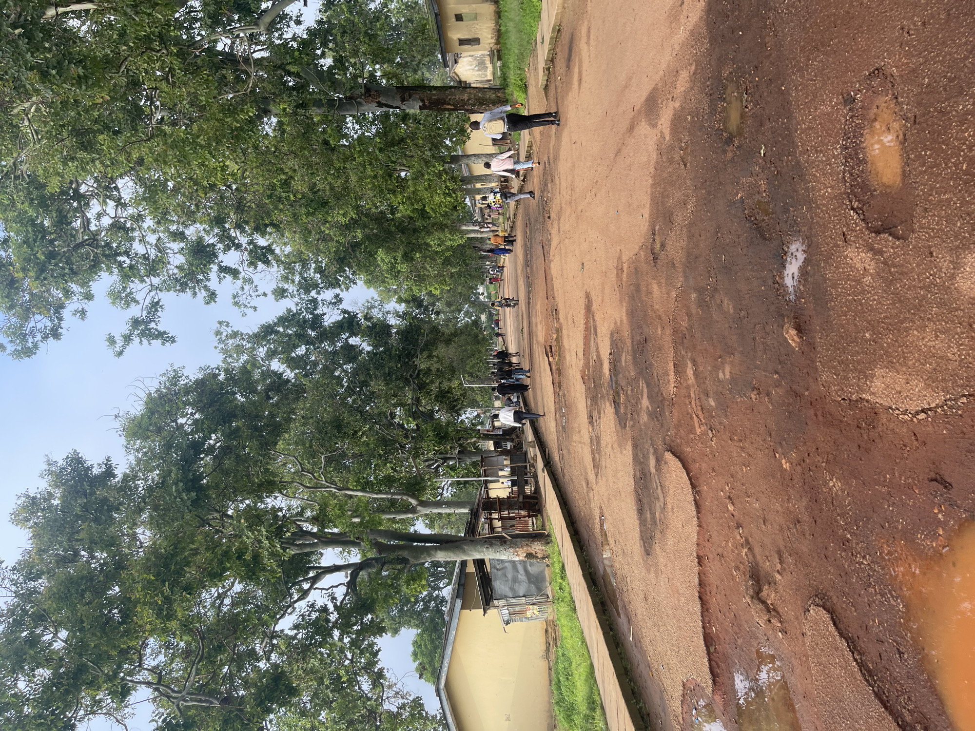 The image depicts a wide, unpaved dirt road in Rafin Par, Nigeria, lined with mature trees on both sides. The road surface is heavily eroded, with numerous large potholes and puddles of muddy water scattered throughout, particularly in the foreground. On the left side of the road, there are yellow buildings and makeshift stalls or structures constructed with corrugated metal and wood. On the right side, more yellow buildings are visible, set back from the road. Several people are walking along the road and the adjacent raised footpath. In the mid-ground, a person on a motorcycle is visible. The sky is overcast, suggesting a cloudy day. The scene captures a typical rural or semi-urban setting with infrastructure in a state of disrepair.