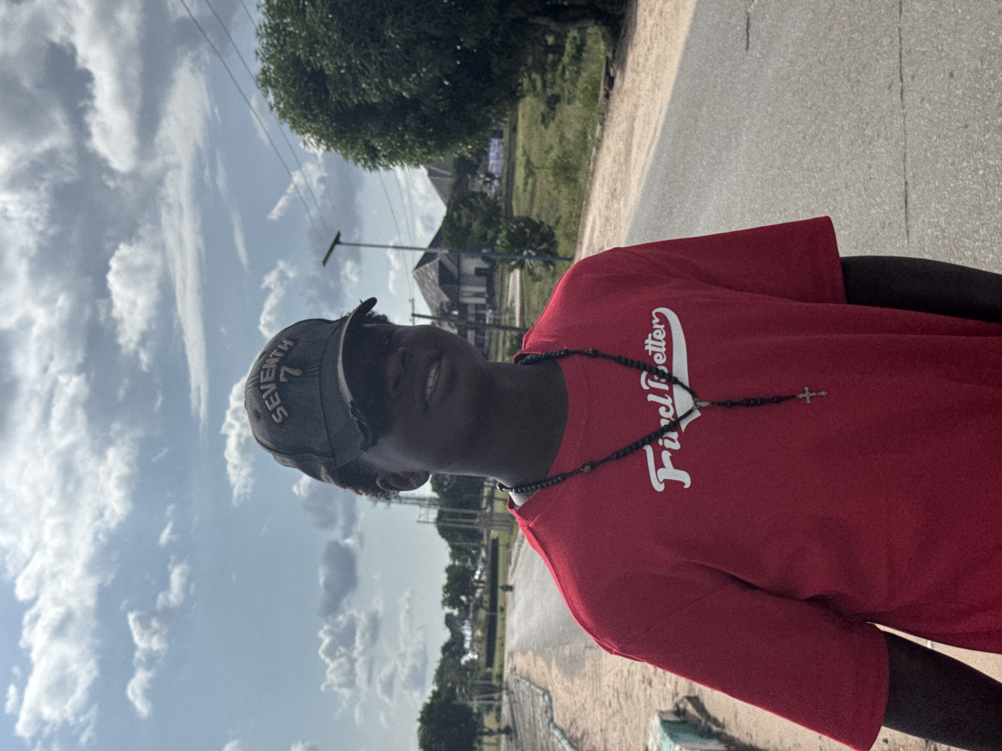 The image captures a young individual, likely male, outdoors during the day in Ihiagwa, Nigeria. The person is positioned in the foreground, looking directly at the camera with a faint smile. They are wearing a red t-shirt and a dark baseball cap with the word "SEVENTH" partially visible on its front. A dark, beaded rosary-style necklace with a small silver cross pendant hangs around their neck. The red t-shirt features white text across the chest, which appears to read "Fired Better" or a similar phrase.
The background shows a bright, partly cloudy sky above a landscape with lush green trees and foliage. A paved road or path runs alongside the person. In the mid-ground, there are utility poles and power lines, with several simple buildings or houses nestled among the trees in the distance. The lighting suggests a clear day, possibly late morning or afternoon. The overall scene conveys a casual and serene outdoor moment.