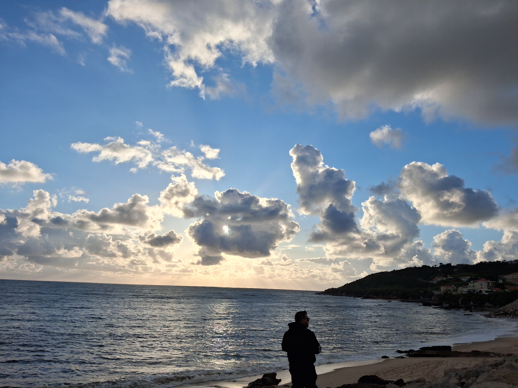 The image depicts a coastal scene in Figueira da Foz, Portugal. The sky is predominantly blue with scattered cumulus clouds, some appearing dark and dense, and sunlight breaking through the gaps. Below, the Atlantic Ocean stretches towards the horizon, reflecting the light. The waves are visible near the shoreline, where the water meets a sandy beach.
A person stands on the beach facing the ocean. A hillside with vegetation and some buildings is visible in the background, along the coastline. The sun is setting over the water.