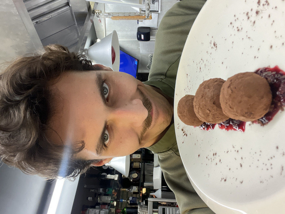 The image shows a man in what appears to be a professional kitchen setting, presenting a meticulously prepared dessert. The image is rotated 90 degrees to the left, making the man's head and the background appear sideways.
The central figure is a man with dark, curly or wavy brown hair and striking light-colored eyes, possibly green or blue. He has a neatly trimmed mustache and some stubble on his chin, forming a light beard. He is looking directly at the viewer with a slight smile or smirk, conveying a sense of pride or satisfaction. He is wearing an olive green or khaki-colored short-sleeved shirt, possibly a chef's jacket.
To the right of the man, and occupying a significant portion of the image, is a white round plate holding three identical, sphere-shaped dessert items. These items are brown, likely chocolate truffles or similar confectioneries, and appear to be dusted with cocoa powder or a fine spice. They are arranged in a linear fashion on a bed of dark red or purplish compote or sauce, which provides a contrasting color and texture. The plate itself is speckled with small dots of what looks like cocoa powder, adding to the aesthetic presentation of the dish.
The background reveals elements of a commercial kitchen. Above the man's head, there are stainless steel surfaces, possibly exhaust hoods or shelving. Further back, there are shelves filled with numerous bottles, suggesting a bar area or a well-stocked pantry. A white, bell-shaped light fixture is visible on the left side of the background. A blue screen or monitor can be glimpsed in the distance, behind the light fixture. The overall lighting is bright and even, typical of an indoor, professional environment.
The image captures a moment of culinary creation and presentation, with the chef proudly showcasing his dessert.