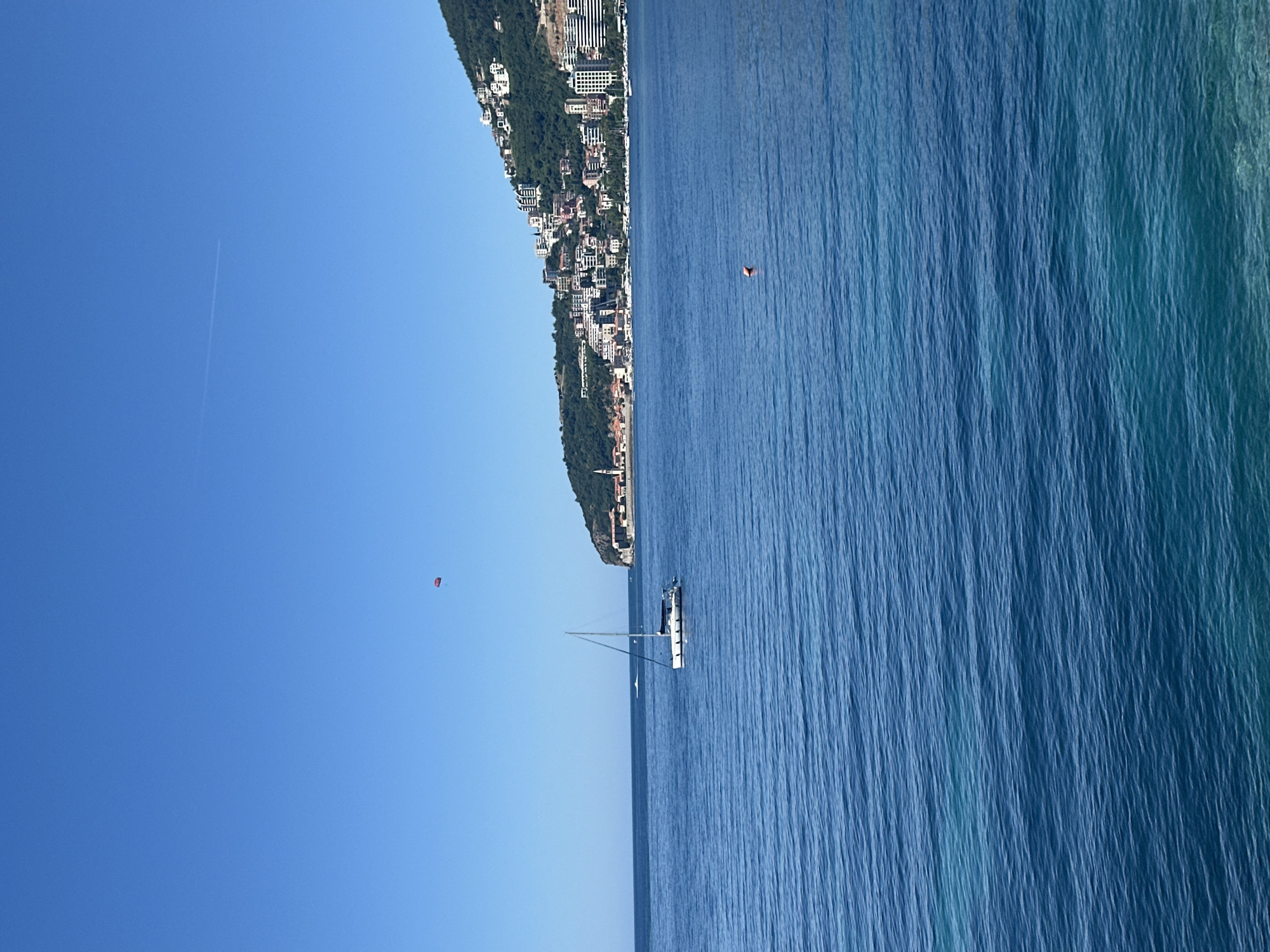 The image shows a seascape in Budva, Montenegro. The scene is dominated by the blue hues of the sea and sky. A sailboat is visible in the water, and a town with buildings on a hillside is located along the coastline. A person is swimming in the sea near the buildings. A red object that may be a parasail is visible in the sky. There is also a contrail left by an airplane visible in the sky.