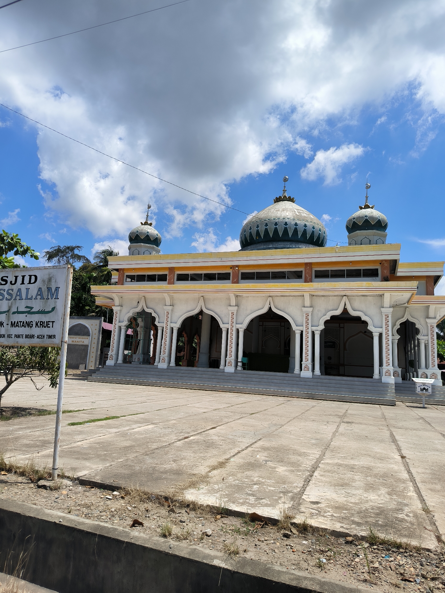 The image shows a mosque called Masjid Assalam in Matangkruet, Indonesia. The mosque is a one-story building with a large central dome and two smaller domes on either side. The building has an arched entrance with white pillars, and there are windows along the top. In front of the mosque is a paved courtyard. A sign with the name of the mosque is visible on the left side of the image. The sky is blue with some clouds. There is a small sign on the right of the entrance.