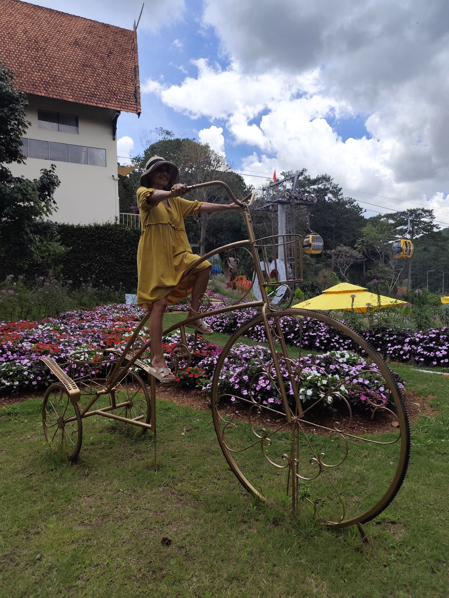 A woman is sitting on an ornamental, penny-farthing-style bicycle in a garden in Da Lat, Vietnam. She is wearing a yellow dress, sandals, and a hat, and is holding the handlebars. The bicycle is gold-colored and stands amidst a bed of colorful flowers. Behind the bicycle are yellow umbrellas, other vegetation, and ski lift lines with gondolas. A building with a red-tiled roof is visible to the left. The sky is partly cloudy.