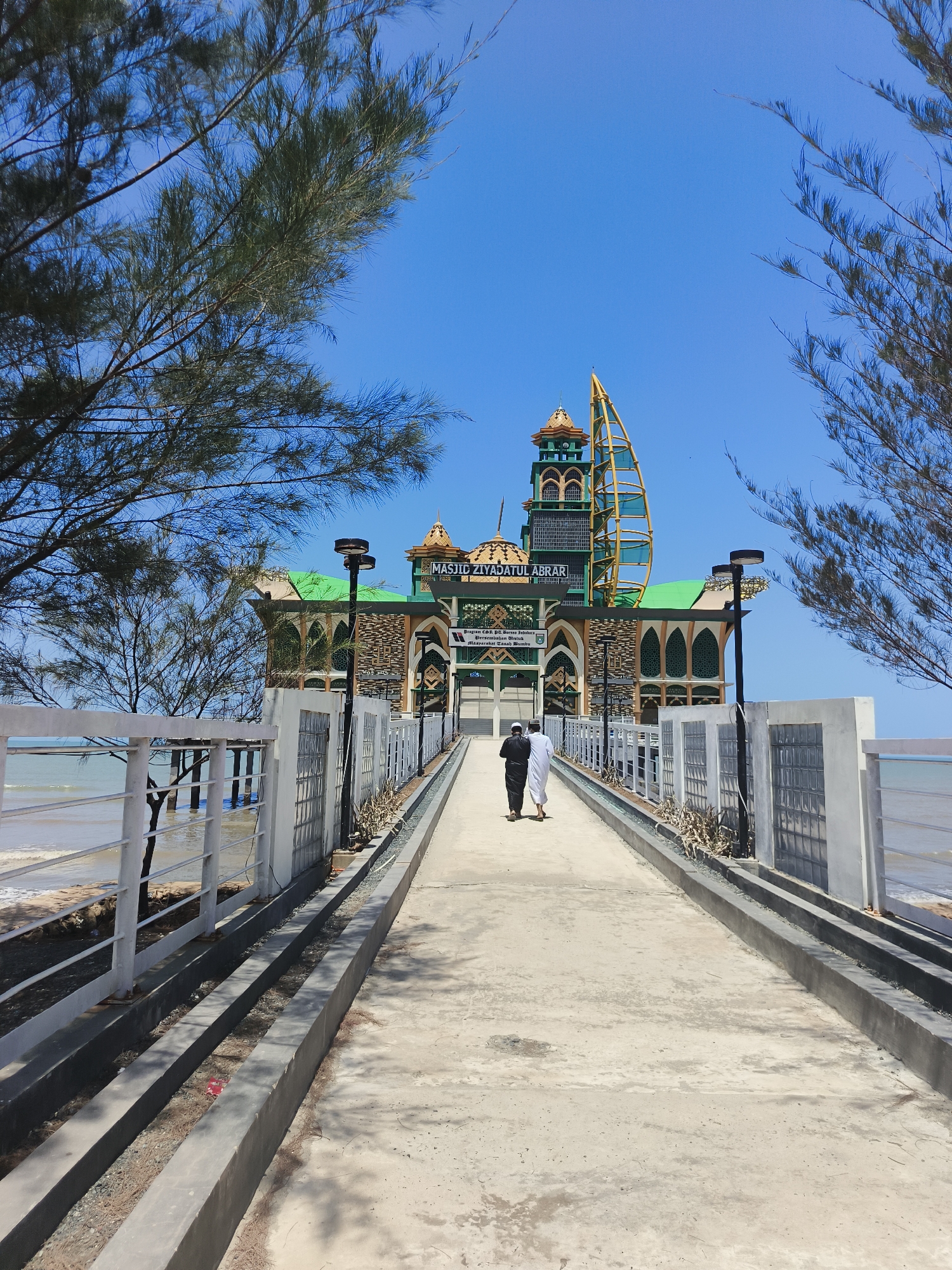 In Beringin, Indonesia, a path leads toward the distinctive Masjid Ziyadatul Abrār, a mosque situated on the coast under a clear blue sky. The mosque features a green roof, colorful ornate architecture with prominent minarets, and a unique yellow sail-like structure on its right side. The path is lined with white railings and a concrete walkway, with the ocean and a sandy shore visible to the left. Two individuals, one in a black garment and the other in white, walk away from the viewer towards the mosque. Pine trees frame the scene on either side, their branches partially obscuring the sky and the upper portions of the mosque. The overall environment is sunny and bright.