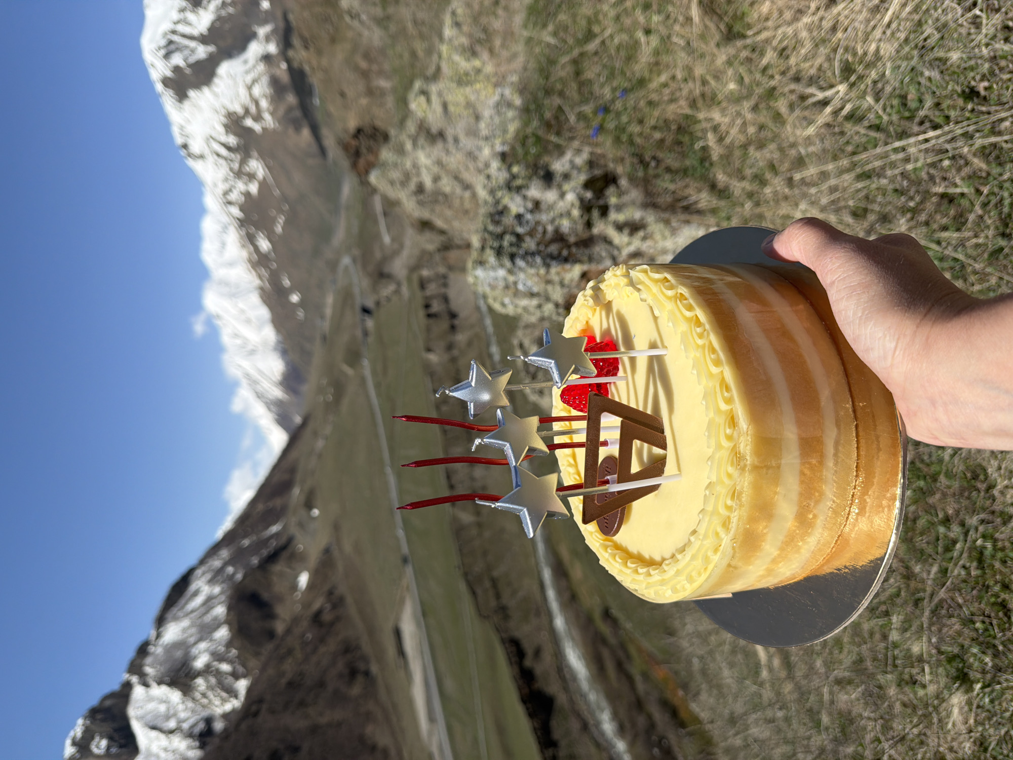 A person holds a cake with star-shaped decorations and candles against a backdrop of mountainous terrain. The cake is round, appears to be multi-layered, and has a creamy frosting. It is positioned on a silver cake board. The background reveals a mountainous landscape with snow-capped peaks visible in the distance. The terrain in the mid-ground is grassy and appears to be dry, with the base of the mountain appearing to be rocky. The sky is partially clear with areas of blue sky and some cloud cover. The scene is located in Pkhelshe, Georgia, as evidenced by the distinctive mountain landscape.