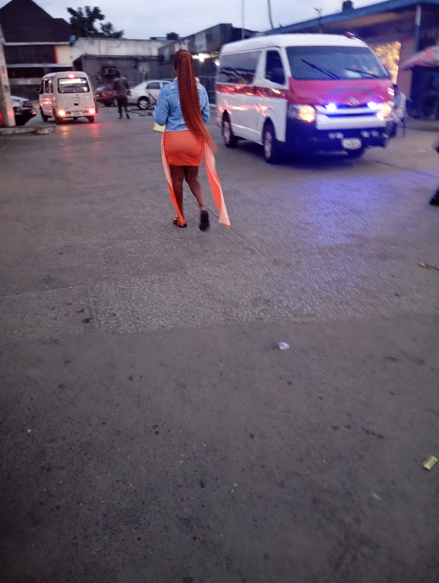 The image shows a street scene in Port Harcourt, Nigeria, likely in the evening or at dusk. In the foreground, a woman with long, braided red hair walks away from the camera. She is wearing a light blue denim jacket and an orange skirt with a white sash draped over it. Her hair is a striking feature, extending well past her waist.
In the background, a white van with a red front and blue flashing lights is parked, suggesting it might be an emergency vehicle or similar service. To the left of the van, a smaller white van is also visible. Several other vehicles and individuals are present in the scene, but they are less distinct due to the lighting and focus. The ground is paved and appears to be a road or parking area. Buildings line the street in the background, with a mix of dark and lighter structures. The overall atmosphere suggests a busy urban environment.