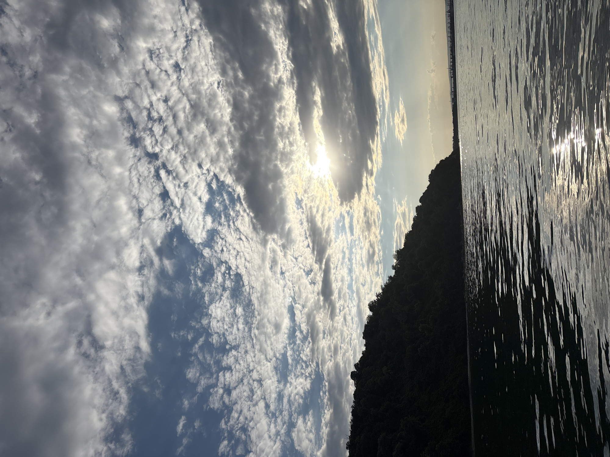 The image depicts a body of water with a tree-covered hillside silhouetted against a dramatic sky. The sun is partially obscured by clouds, casting rays of light across the water, creating a shimmering effect. The clouds themselves are a mix of dark gray and bright white, indicating a dynamic weather pattern. In the distance, a faint industrial landscape with cranes is visible on the horizon. The overall mood is one of natural beauty and the power of the elements.