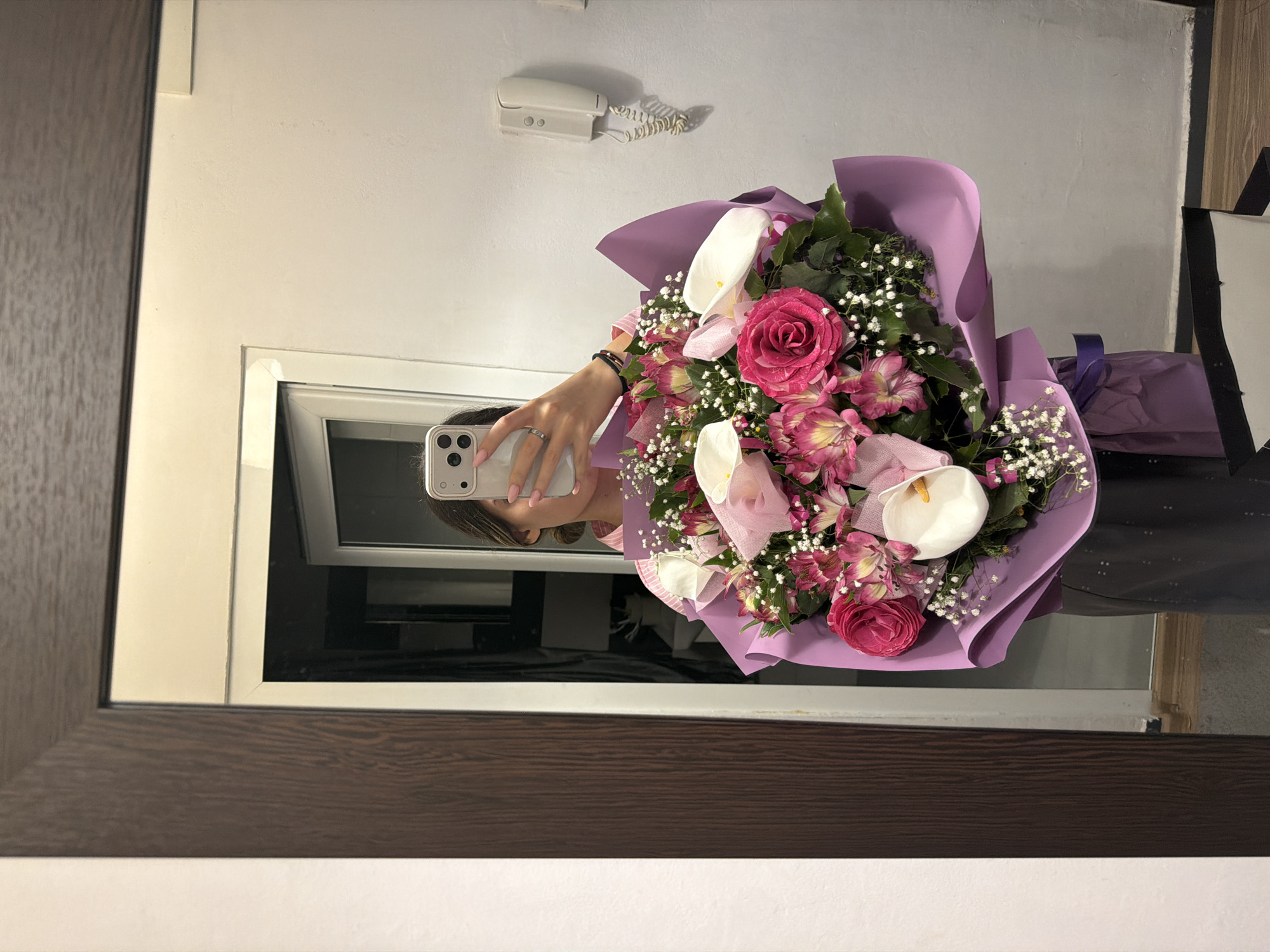 A person is holding a bouquet of pink and white flowers wrapped in purple paper, taking a picture of themselves in a mirror. The mirror has a dark brown wooden frame. The reflection in the mirror shows a white wall, a white phone on the wall, and a window. The location cannot be determined.
