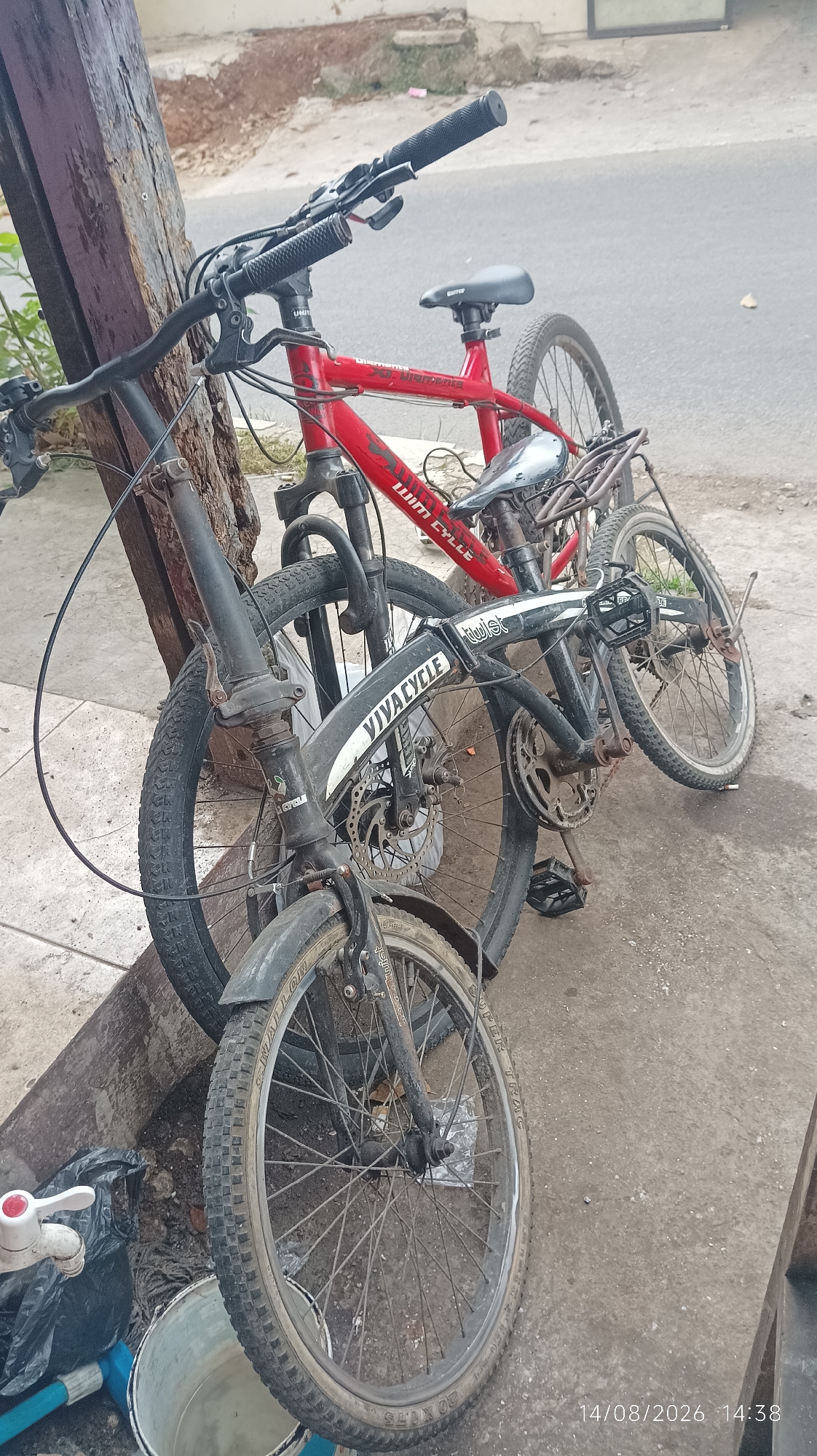 Two bicycles are parked next to a wooden pole on a concrete sidewalk in Tasikmalaya, Indonesia. The bicycle in the foreground is black and folded. It has a disc brake on the front wheel and a tire labeled "VIVA CYCLE". The bicycle behind it is red and has a rear rack. Its tire is labeled "WIM CYCLE". The background shows a street and some vegetation. A white tap with a red handle is attached to a black plastic bag to the left of the bicycles. A bucket with water is next to the bag. The timestamp "14/08/2026 14:38" is visible in the lower right corner.