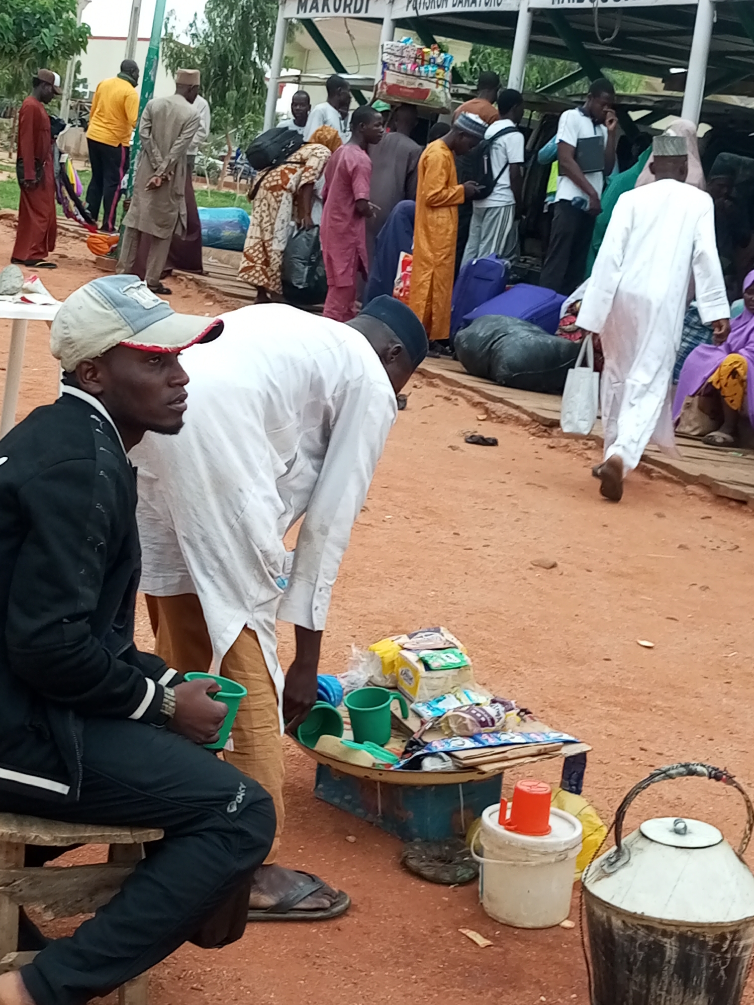 In Gombe, Nigeria, an outdoor scene depicts a bustling market area, possibly a bus station or transit point. The ground is composed of reddish-brown dirt. In the foreground, a man wearing a black jacket and black pants sits on a wooden stool, holding a green plastic cup. Beside him, another man in a white garment and brown pants bends over, reaching towards a tray laden with various packaged goods. A variety of items are spread across the ground, including luggage, buckets, and a large metal pot. In the background, several individuals are visible, some carrying bags and others appearing to be waiting. A structure with signage, partially visible at the top, suggests a commercial establishment. The overall atmosphere is one of activity and movement, typical of a busy public space.