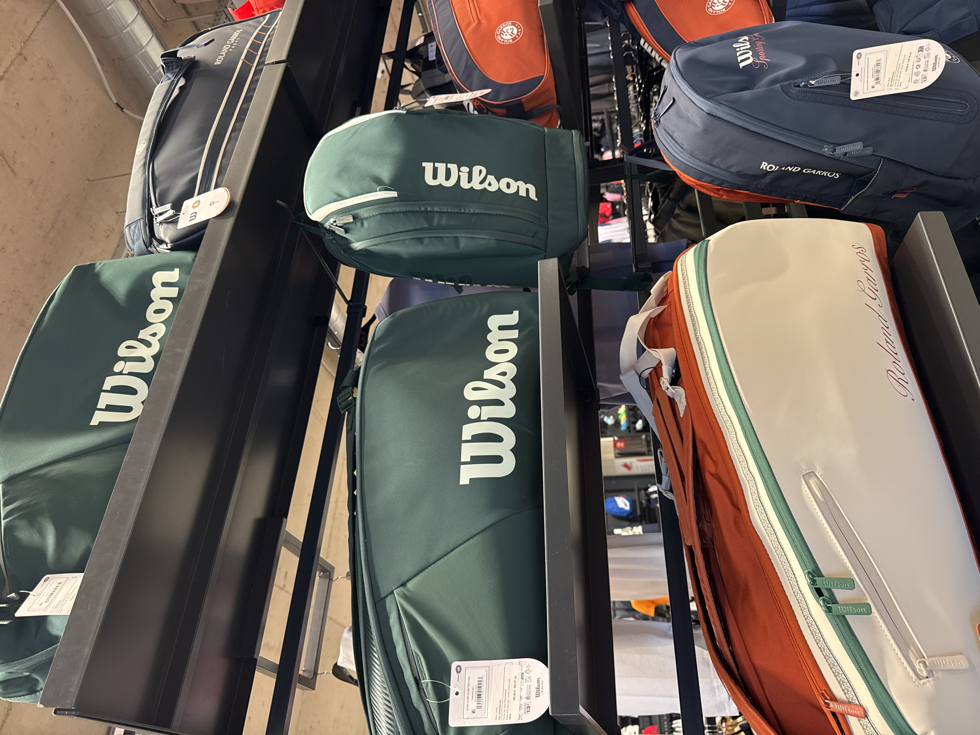 This image captures a retail display of tennis bags, likely within a sports store or pro shop. Several rows of bags are arranged on dark metal shelving units, viewed from a slightly elevated perspective. The bags primarily feature prominent "Wilson" and "Roland Garros" branding, indicating sports equipment associated with the French Open tennis tournament.
The color palette of the bags is dominated by dark forest green, navy blue, and a warm terra cotta or burnt orange, often accented with white or lighter hues. Multiple bags, especially those in dark green, clearly display "Wilson" in large, white block letters. Other bags, particularly the navy and orange ones, feature "ROLAND GARROS" text. One distinct white bag showcases "Roland Garros" in an elegant cursive script. Visible zipper pulls are embossed with the "Wilson" logo.
Product tags are attached to several bags, displaying barcodes, various product information, and "Wilson" branding. A tag on a navy bag also shows part of "Wilson Sporting G." The overall scene suggests an indoor commercial setting, a sports equipment store, located in Sofia, Bulgaria.