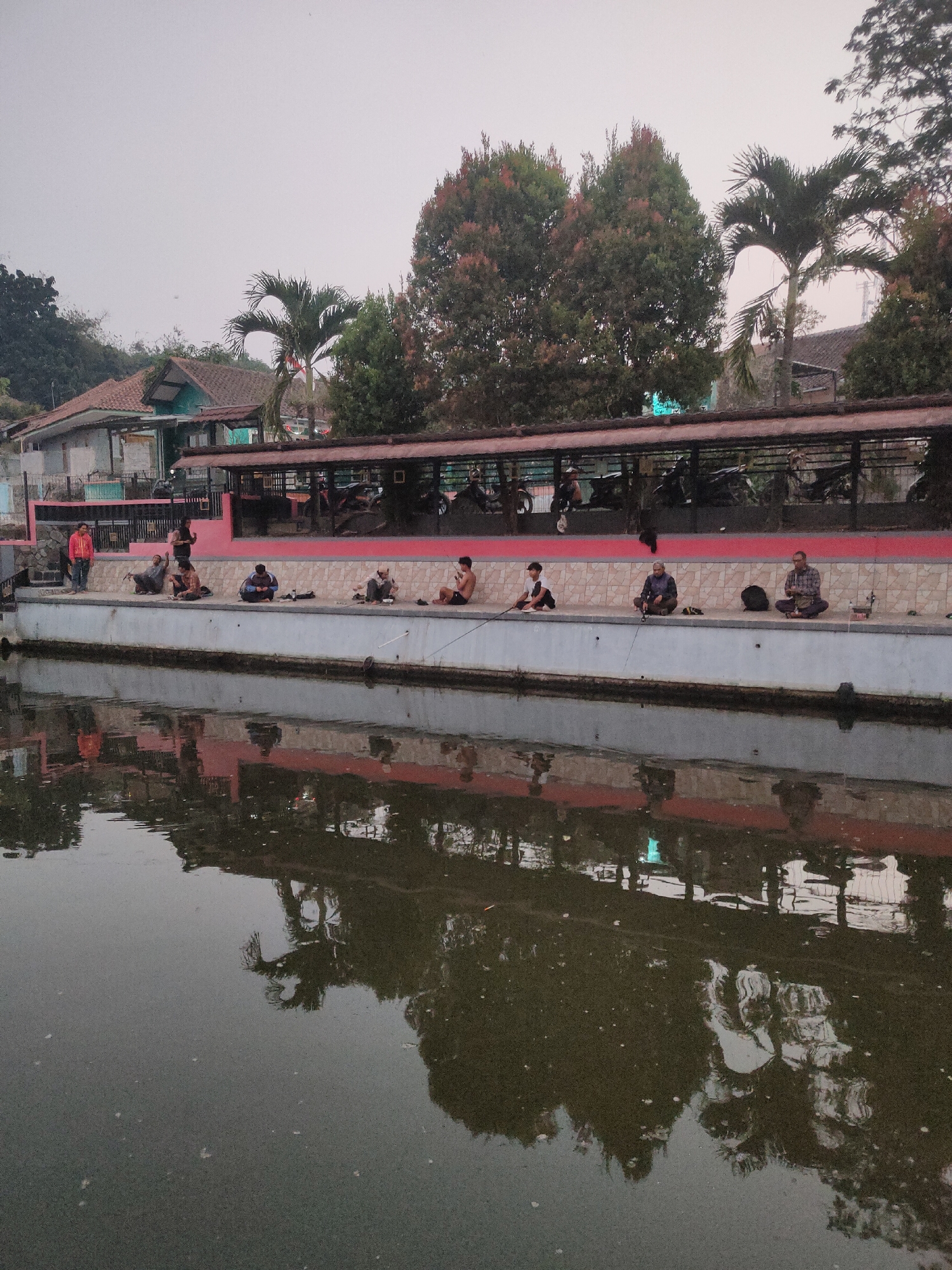 The image captures a serene afternoon scene by a body of water, likely a canal or river, in Sumedang, Indonesia. Several individuals are engaged in fishing from the elevated concrete embankment that borders the water. They are seated, with fishing rods extended into the water, suggesting a leisurely and patient activity. The embankment itself is lined with a decorative tiled wall and a vibrant pink base, suggesting a public or recreational area. Above the embankment, a covered structure with a red roof is visible, possibly a pavilion or market stall, with motorcycles parked beneath it. In the background, a cluster of houses with reddish-brown tiled roofs and some lush greenery, including palm trees, are nestled on a gentle slope, indicating a semi-urban or suburban setting. The water's surface reflects the surrounding environment, including the embankment, the structure, and the trees, adding a sense of depth and tranquility to the scene. The overall atmosphere is peaceful, with the focus on the quiet pursuit of fishing against a backdrop of local Indonesian architecture and natural elements.