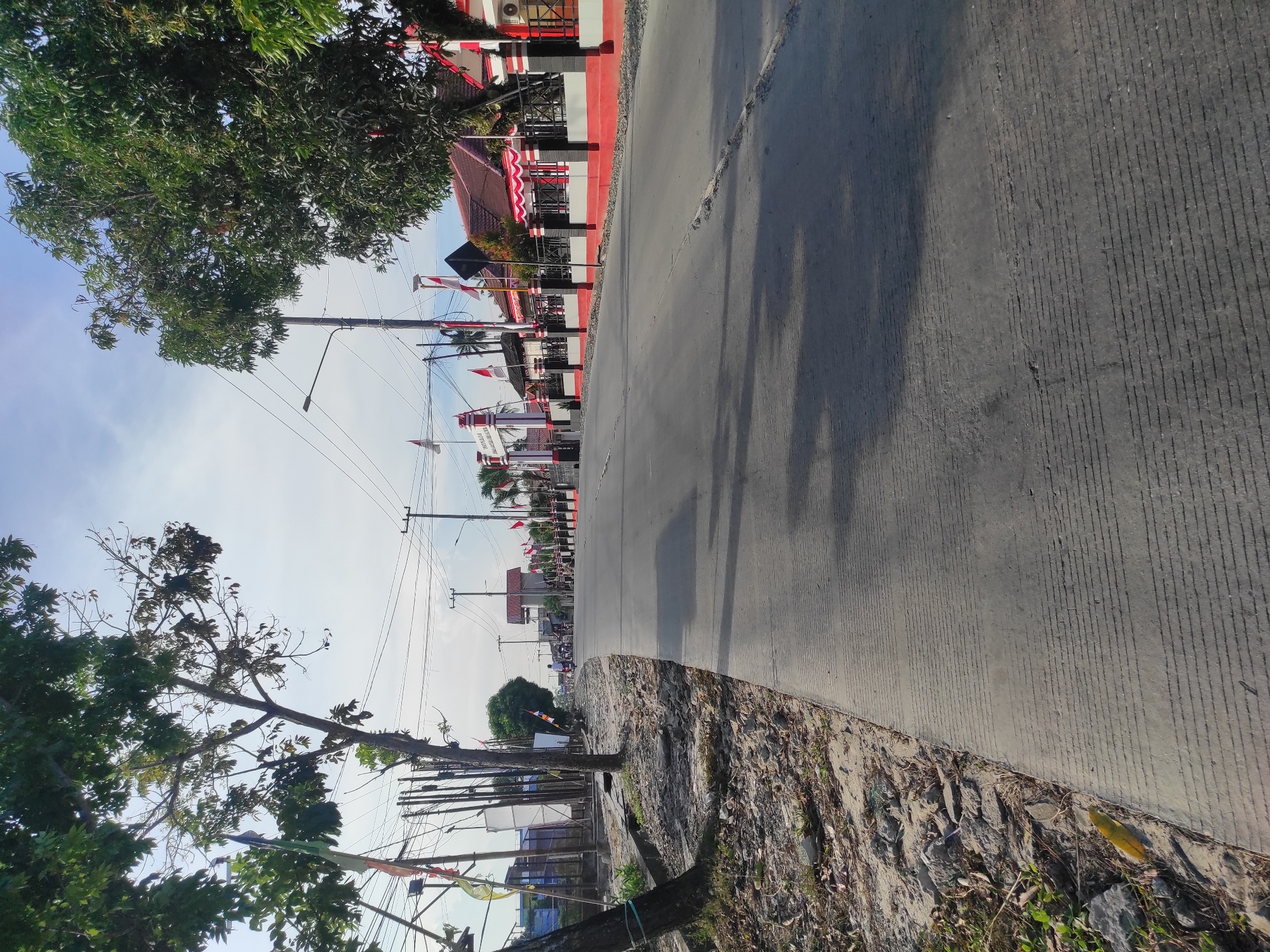 The image depicts a street scene in Muara Jawa Ulu, Indonesia. A paved road occupies the foreground and midground, curving to the right. The road surface is grey concrete with horizontal lines imprinted for texture. To the left of the road, a dirt and gravel embankment is visible, with scattered stones and sparse vegetation. Several trees with green leaves are present on the left side, casting shadows on the road.

On the right side of the road, a building with red tiled roofs and white and red striped awnings is visible. This building appears to be a commercial establishment or a public facility, with a black metal fence running along its front. Several Indonesian flags are displayed prominently, attached to poles along the street and on the building. In the distance, more flags and buildings can be seen, suggesting a populated area.

Several utility poles with numerous wires are present throughout the scene, indicating an urban or semi-urban setting. A few individuals on motorcycles are visible further down the road, heading away from the viewer. The sky is partly cloudy with a light blue hue, and the overall lighting suggests it is daytime, likely morning or late afternoon due to the angle of the shadows.