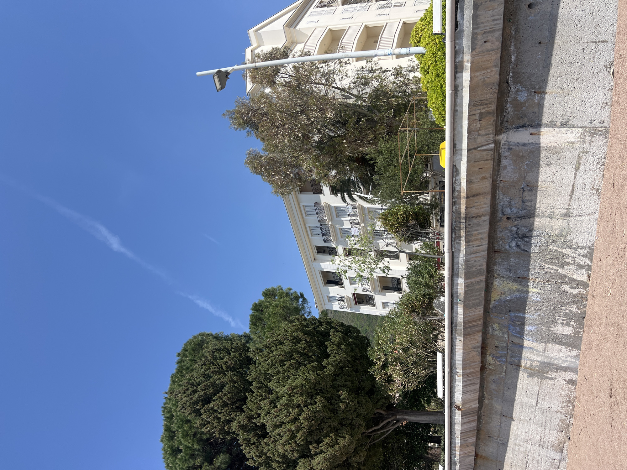 The image displays a sunlit outdoor scene in Petrovac centar, Montenegro, under a clear blue sky. The viewpoint is angled upwards, showcasing a blend of architecture and natural elements.
In the upper part of the frame, a vast expanse of clear blue sky is visible, featuring a thin white contrail stretching diagonally across it. A modern, white street light fixture, attached to a pole, extends from the right side towards the sky.
Below the sky, on the left, are the dense, green canopies of tall trees, possibly cypress or similar evergreens, forming a rich, textured mass of foliage. To the right, a prominent multi-story building, painted white, stands out with numerous balconies featuring dark railings and ornate details. Several white rectangular signs with dark, unreadable text are attached to the balconies, likely indicating properties for rent or sale.
Further to the right and in the middle ground, another cluster of trees with lighter green, deciduous foliage partially obscures the lower sections of the building. A yellowish object, possibly a garbage bin, and a metallic, grid-like structure (perhaps scaffolding or a stand) are nestled among these trees.
The foreground is dominated by a weathered concrete barrier or retaining wall running horizontally across the lower right. This wall exhibits various shades of grey, with visible staining, discoloration, and some light graffiti or paint marks. A grey pipe or railing runs along the top edge of this barrier. The ground immediately adjacent to the barrier is a reddish-brown color, suggesting a path or unpaved area.
The scene depicts a bright, clear day, likely midday or early afternoon, given the strong illumination and shadows. No people or specific activities are visible, presenting a serene, static view of a Mediterranean urban landscape.