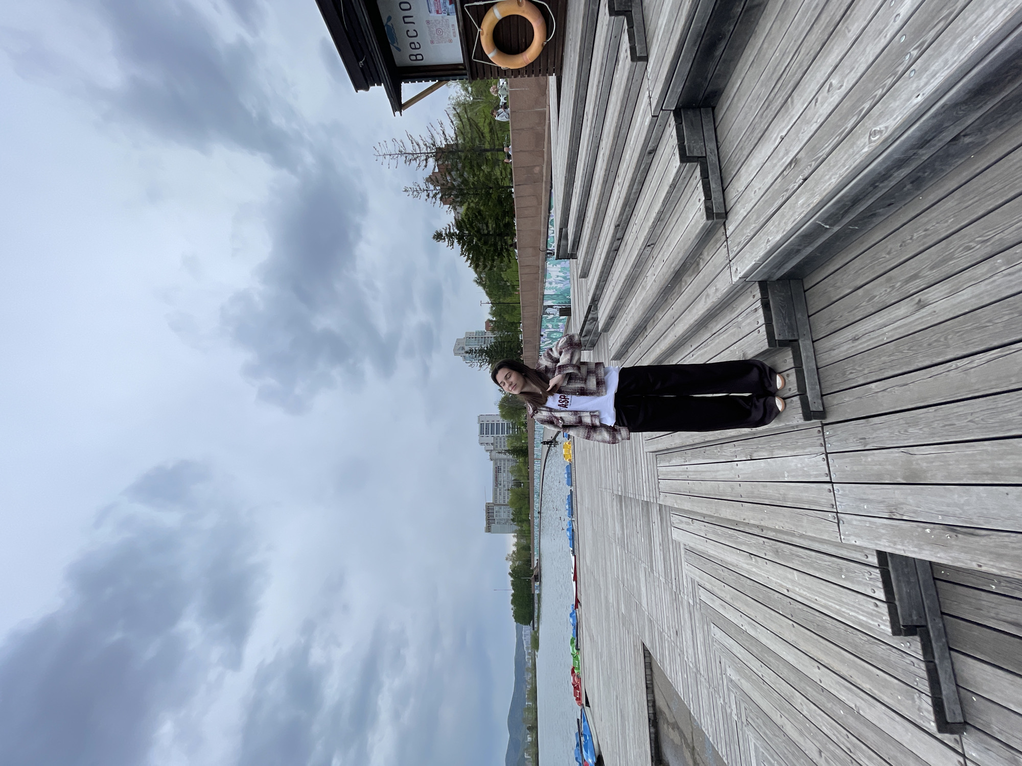 The image shows a young woman standing on a wooden boardwalk by a body of water, with buildings and mountains visible in the background. The sky is overcast. The boardwalk is made of grey wooden planks. The woman is wearing a plaid jacket, a white t-shirt, and black pants. A sign with Cyrillic text and a life preserver are mounted on a structure in the background to the right of the woman. The location is likely in Krasnoyarsk, Russia. There are paddle boats near the water.