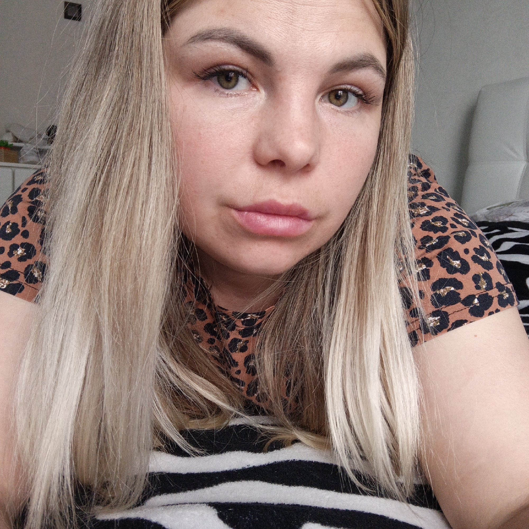 A close-up shot of a woman with long, blonde hair, wearing a brown and black leopard-print shirt. She is lying on a black and white striped surface, possibly a blanket or bedding. The location is Unknown, Unknown.