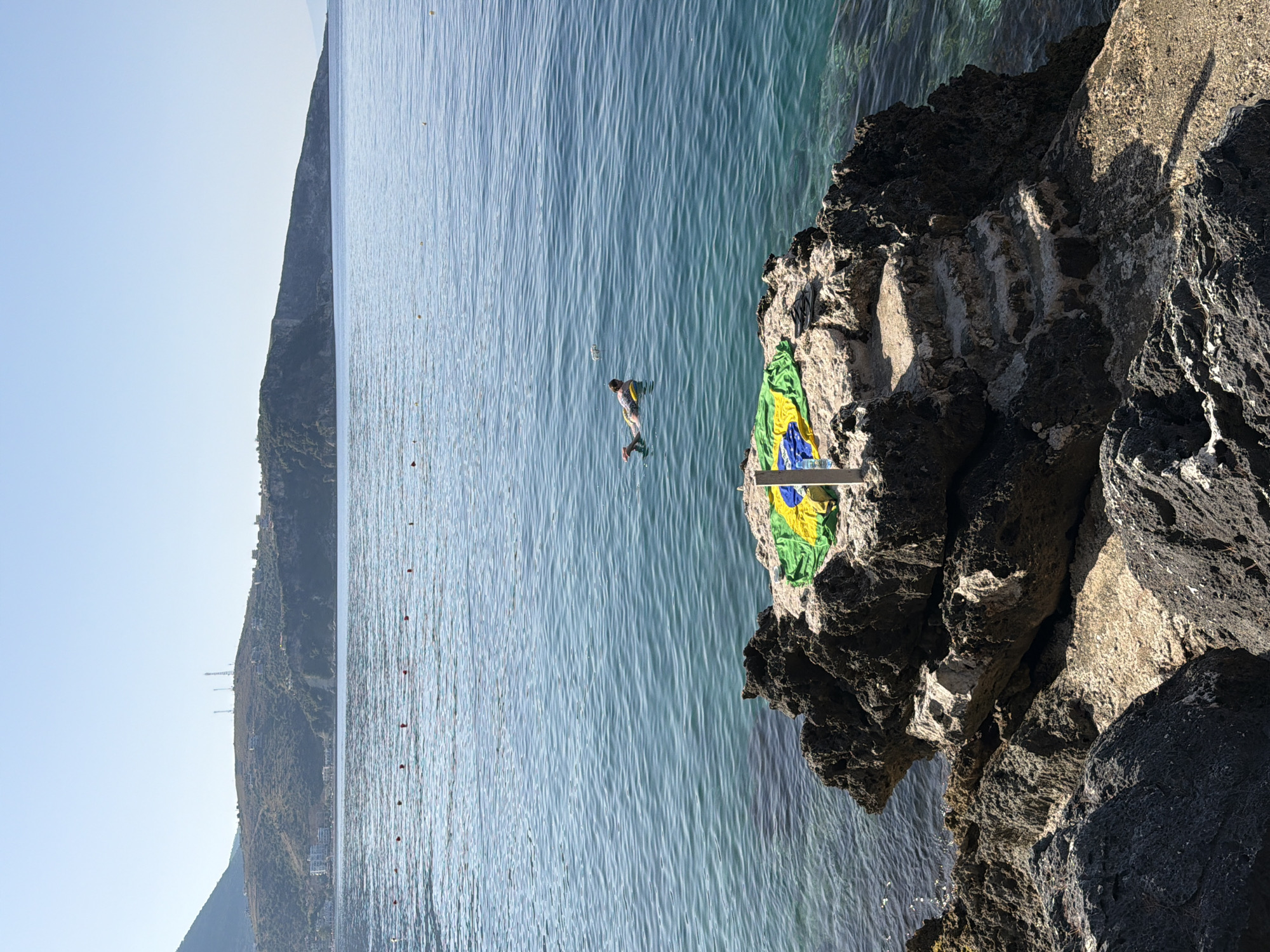A person is swimming on a yellow inflatable ring in the clear turquoise waters of the Ionian Sea, near the city of Himarë, Albania. In the foreground, a rocky outcrop juts into the sea, and on its flat top, a Brazilian flag is laid out. Steps are carved into the rock, leading down towards the water. In the background, a verdant hillside dotted with buildings and topped with communication towers overlooks the sea and the coastline. The sky is clear and blue.