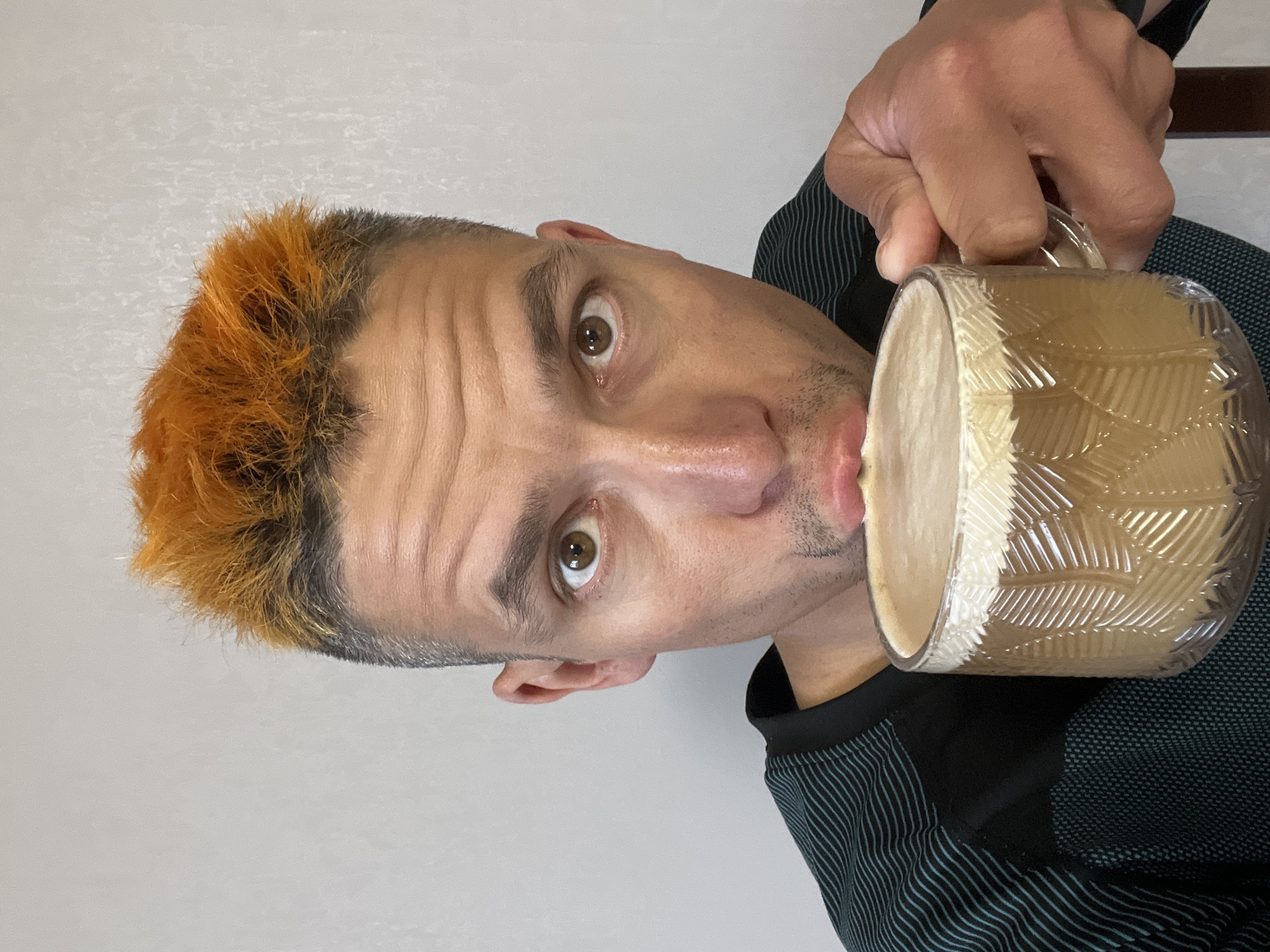 A man with short hair dyed orange on top is seen holding a large, patterned glass mug filled with a frothy beige liquid, possibly coffee or latte, to his lips. He is looking directly at the camera with wide eyes. He's wearing a dark shirt with light blue vertical stripes on the sleeves. The background is a plain, light-colored wall. The image is rotated counter-clockwise. There is no evidence to suggest the location is Chaikovsky, Russia.