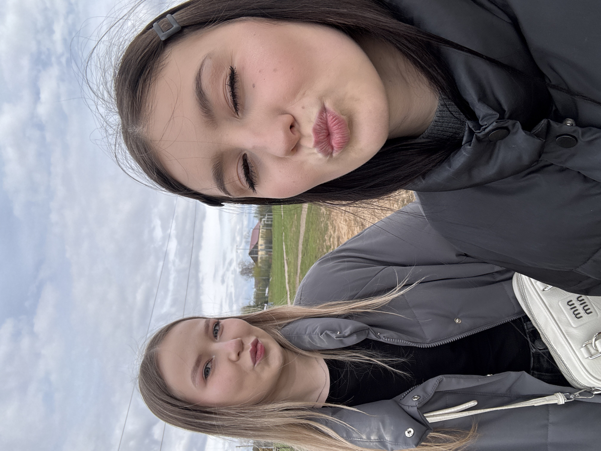 Two young women are featured in a vertical selfie, taken outdoors during daytime under an overcast, cloudy sky. The woman in the foreground has dark, long hair, secured with a dark hair clip, and poses with her eyes closed and lips pursed in a playful "kiss" expression. She wears a black puffer jacket over a dark top. Slightly behind her and to the right is another woman with long, light-colored hair, who is looking towards the camera with a subtle smile. She is dressed in a grey puffer jacket over a black top and carries a light-colored handbag that prominently displays the text "Miu Miu". The background shows a green, grassy field extending into the distance, with some power lines overhead and faint outlines of buildings, possibly houses, on the horizon. The setting suggests a rural or semi-rural location in Фоки, Russia.
