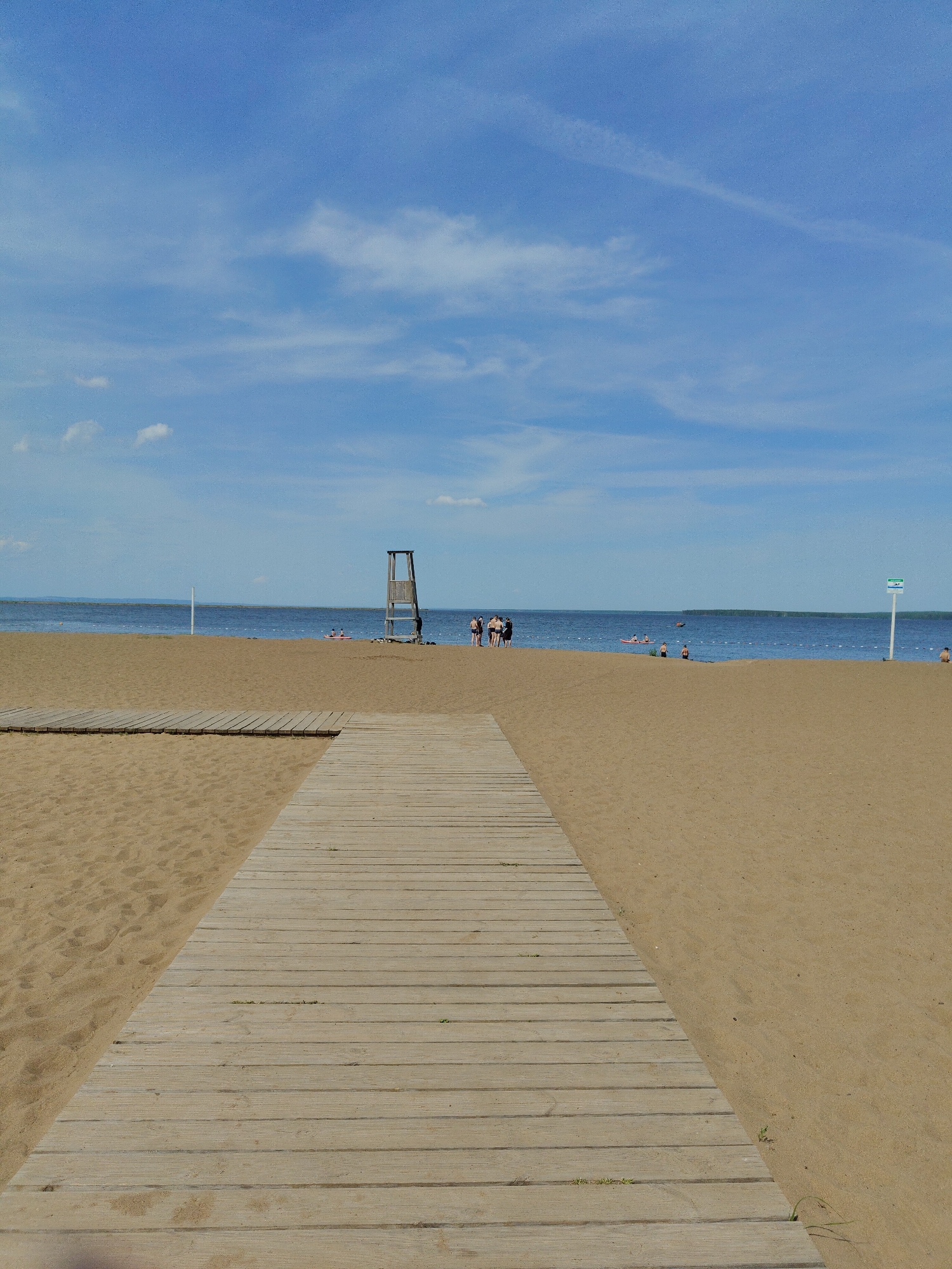 A wooden walkway extends towards a sandy beach bordered by a body of water, likely a lake or ocean, under a bright blue sky with scattered clouds. In the background, several individuals are visible on the beach and in the water. A lifeguard tower is positioned near the center of the frame. A sign stands to the right of the lifeguard tower. The location appears to be a public beach area.