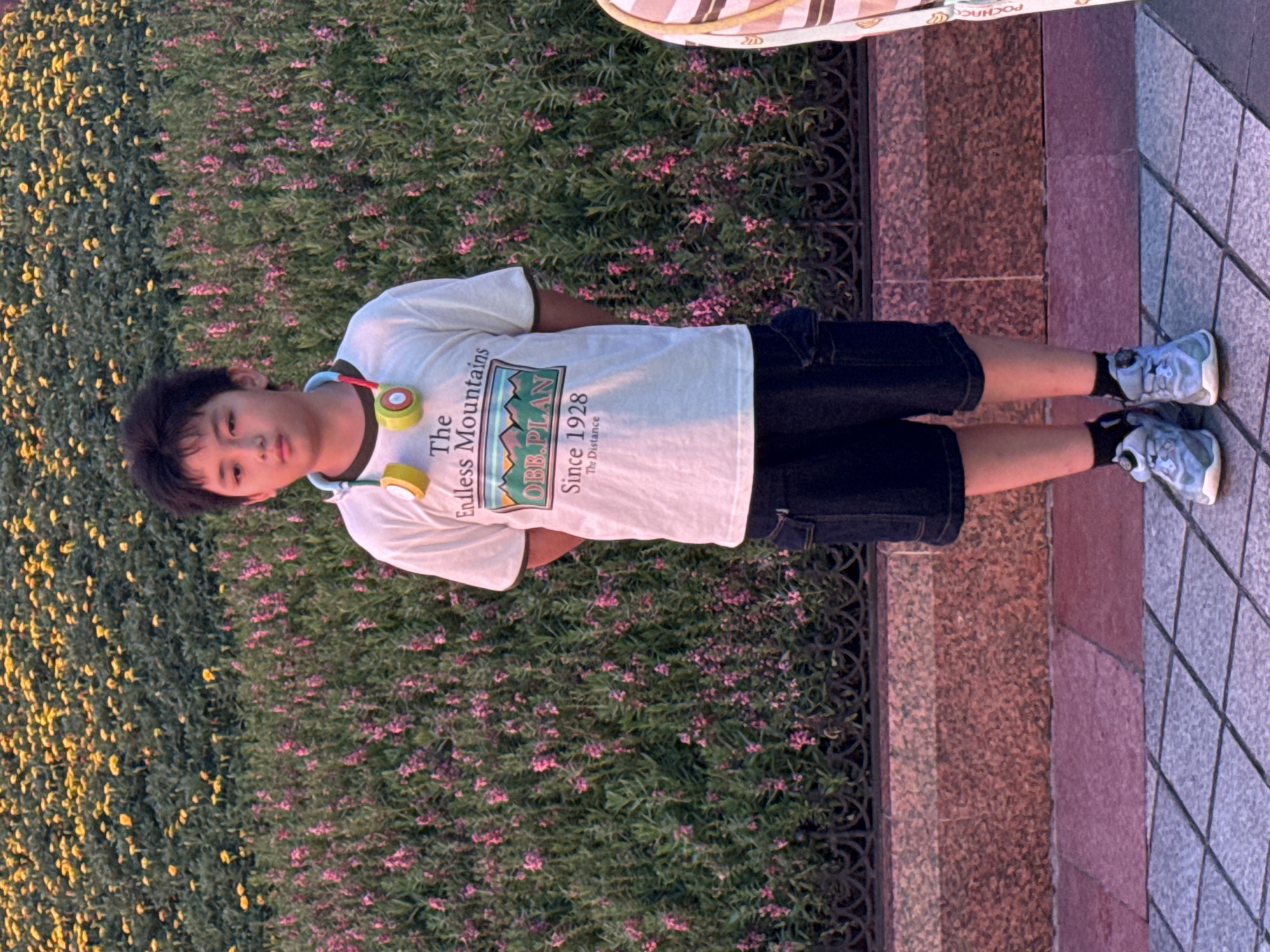 A young East Asian boy stands in front of a dense backdrop of green bushes dotted with small pink and yellow flowers. The boy is facing the camera, his hands clasped behind his back. He has dark hair and is wearing a white t-shirt with a graphic print on the front, dark shorts, black socks, and light blue sneakers. A pair of headphones with yellow earcups and red and green accents rests around his neck. Behind him, a decorative black metal fence separates the flower bushes from a textured stone wall. The ground in front of the wall is paved with square tiles. The lighting suggests it is late afternoon or early evening, with a warm glow. This scene is located in Yuzhong District, China.