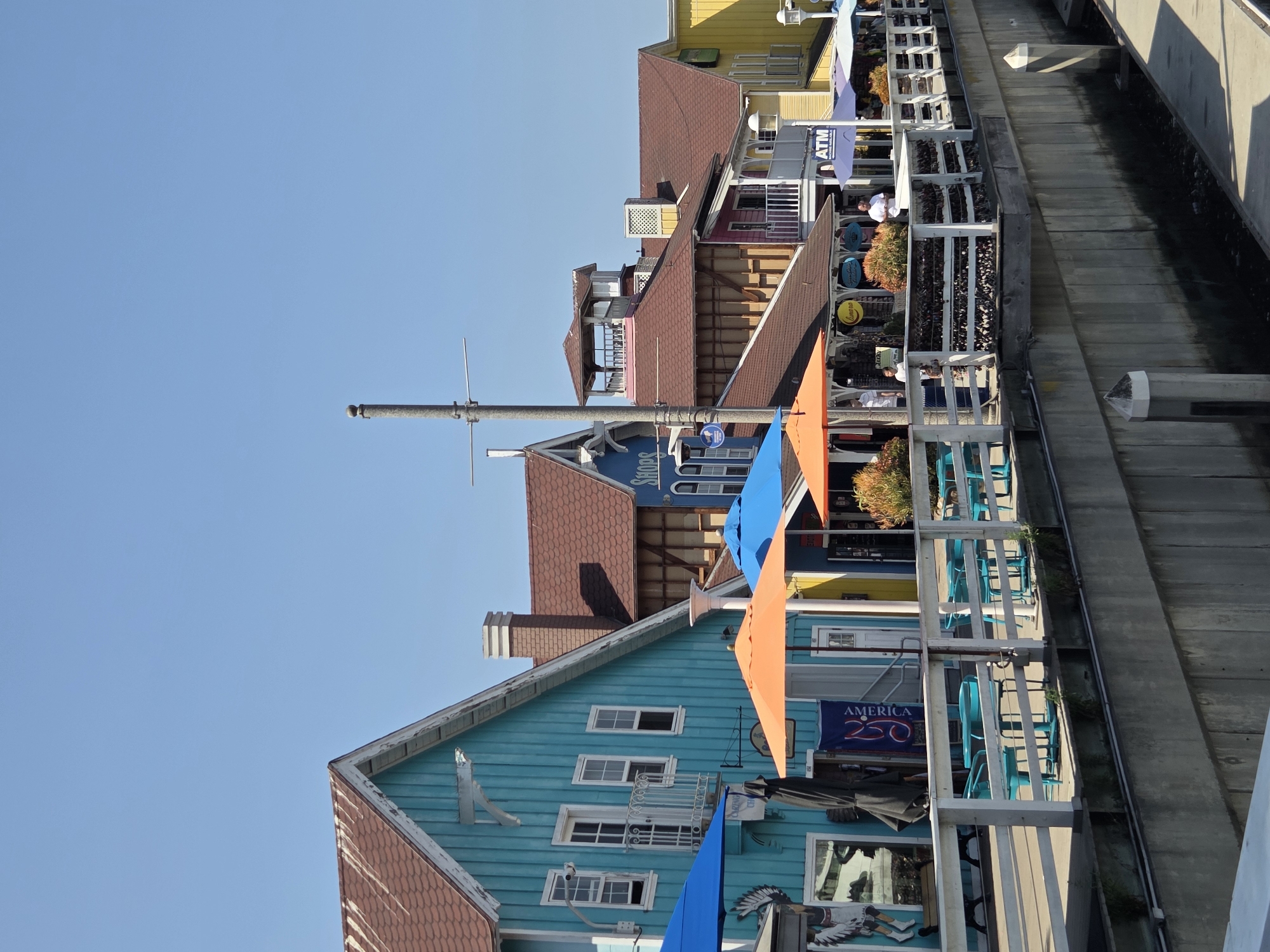 The image depicts a bright, sunny day in Long Beach, California, featuring a row of colorful waterfront buildings. The architecture is varied, with houses painted in vibrant hues of turquoise, yellow, and pink, topped with reddish-brown roofs. Several buildings have decorative trim and balconies. In the foreground, a white railing borders a walkway lined with teal outdoor tables and chairs. Umbrellas, in shades of orange and blue, provide shade over some of the seating areas. A few people are visible on the walkway, some appearing to be shoppers or diners. The buildings are situated along a waterfront, with a concrete seawall in the lower part of the image, suggesting a harbor or canal. Signs on the buildings indicate businesses, including "SHOPS" and "ATM." The sky is clear and blue, emphasizing the sunny weather.