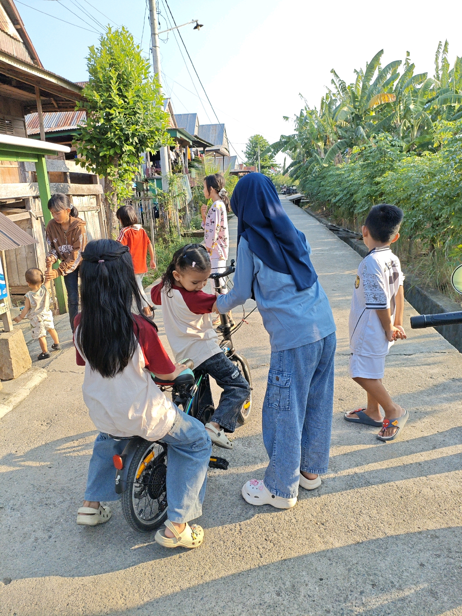 In Padduppa, Indonesia, a group of children and one adult are gathered on a paved street lined with houses and lush banana trees. The scene appears to be captured during daylight hours, with warm sunlight casting long shadows.
In the foreground, two girls are interacting with a bicycle. One girl, with long dark hair, is seated on the bicycle, facing away from the camera. She is wearing a light-colored top with red sleeves and blue jeans. Her feet are adorned with light-colored, possibly croc-style shoes. The other girl, facing the bicycle and partially obscured by it, is wearing a blue headscarf and a light blue long-sleeved shirt with blue jeans. She is reaching towards the bicycle, seemingly engaged with the first girl or the bike itself.
Further in the background, a young boy in a white t-shirt and white shorts stands with his hands behind his back, looking towards the right side of the frame. He is wearing dark blue flip-flops. To the left of the bicycle, an adult woman in a brown top and dark pants is holding the hand of a young child. The child is dressed in a light-colored sleeveless shirt and shorts and is walking towards the left. Behind them, another child in a red shirt is walking away from the camera. A girl in a pink patterned dress is standing further down the street, looking to her left.
The environment includes modest wooden houses with corrugated metal roofs on the left side of the street and dense vegetation, including banana trees, on the right. A utility pole with wires is visible in the background, suggesting the presence of electricity. The road surface is rough and textured. The overall impression is a snapshot of a community gathering on a sunny day in a tropical setting.