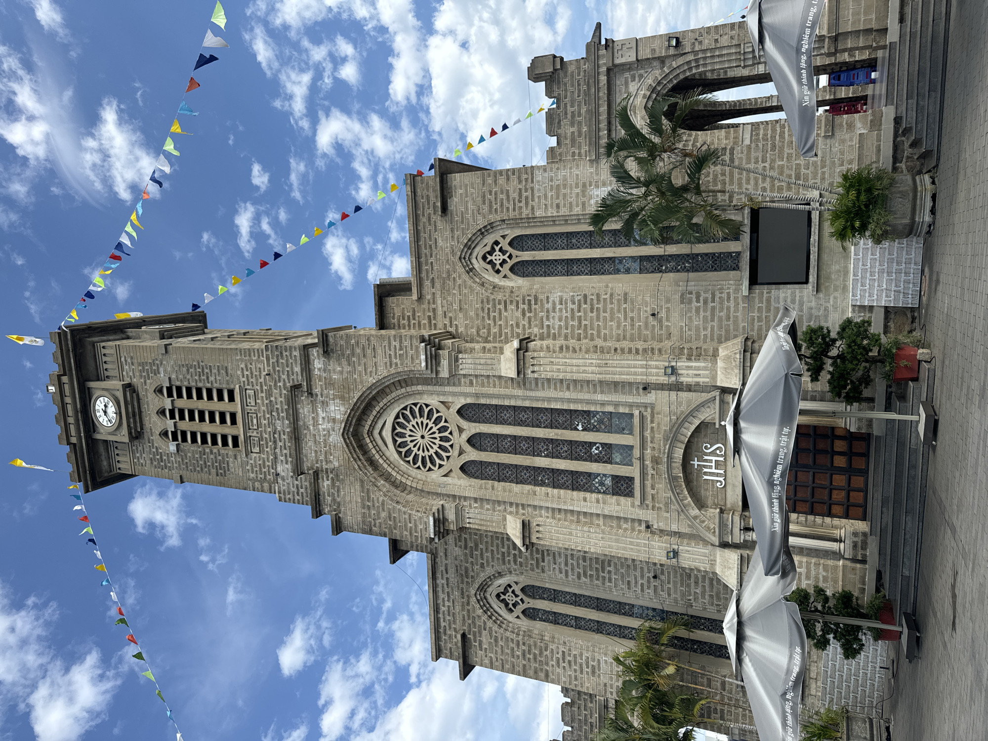 The image captures the imposing facade of the Nha Trang Cathedral, a Roman Catholic cathedral located in the city of Nha Trang, Vietnam. The cathedral is constructed of stone with gothic architectural elements, featuring a tall bell tower with a clock face, arched windows, and a rose window above the main entrance. Colorful triangular flags are strung across the sky above the tower, suggesting a festive occasion. In front of the cathedral, two large grey umbrellas provide shade, with text in Vietnamese visible on their surface. Steps lead up to a large wooden door, above which the monogram "JHS" is mounted. Palm trees and potted plants are visible on either side of the entrance. The sky is blue with scattered white clouds. The overall scene depicts a grand religious building under a bright sky, possibly during a celebration or event.