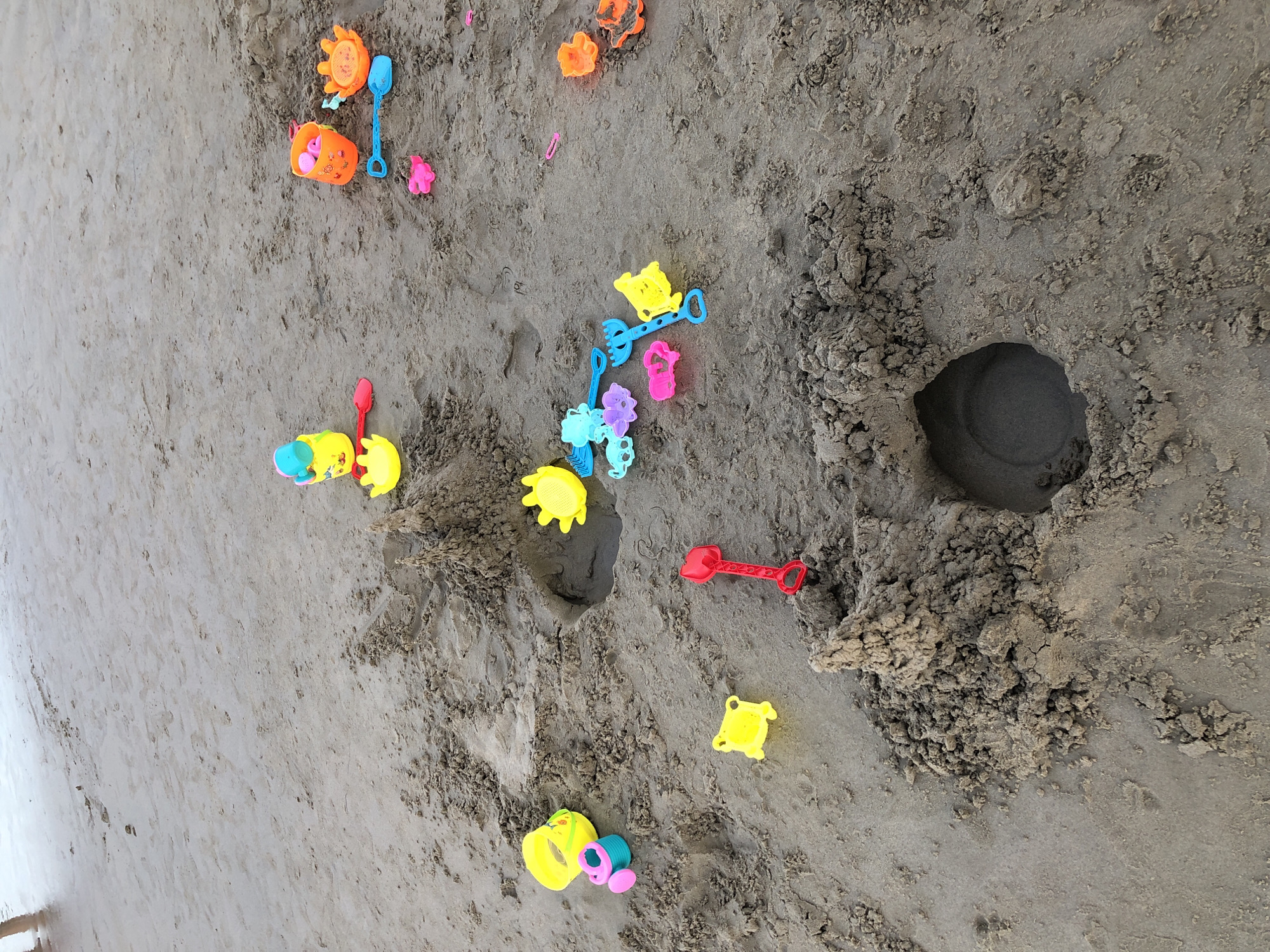 The image shows a collection of colorful plastic beach toys scattered on a sandy beach. There are buckets, shovels, molds shaped like animals, and a small watering can. The sand is damp and textured, with several small piles and indentations, suggesting recent play. In the background, the edge of the water can be seen, with faint footprints leading towards it. The scene is likely located on a beach in Đà Nẵng, Vietnam, a popular coastal city known for its sandy shores and family-friendly atmosphere.
