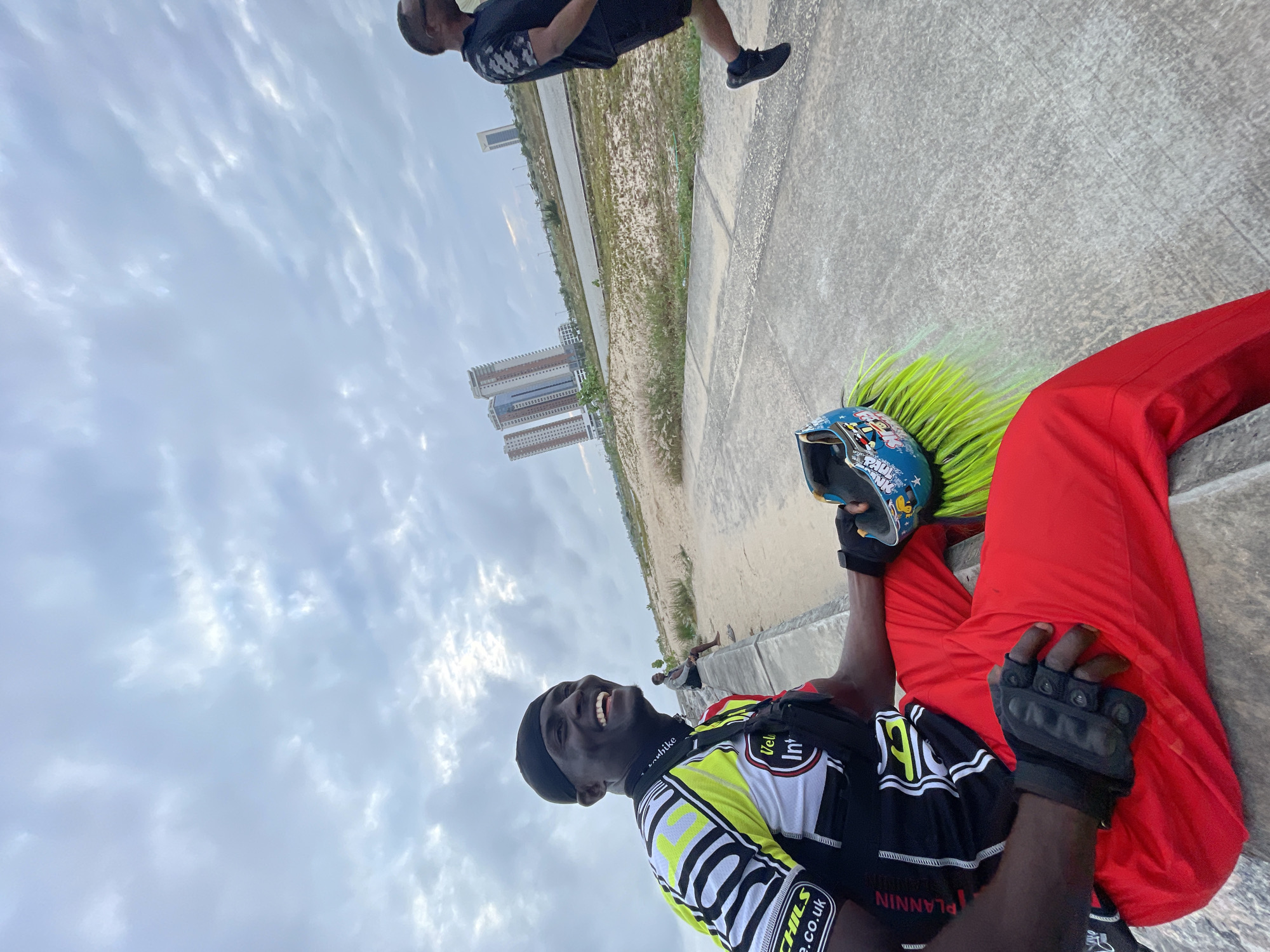 The image depicts an outdoor scene in Inupa Kekere, Nigeria, under a cloudy sky. A Black man in a cycling jersey with yellow, black, and white patterns and bright red pants is sitting on a concrete ledge, smiling broadly. He is wearing a black skullcap and black cycling gloves. A blue helmet with colorful decals and bright green hair-like streamers is placed on his lap. Another man, dressed in a black t-shirt and shorts, is jogging on a paved path in the background to the right. In the distance, several modern high-rise buildings are visible, with a sandy, sparsely vegetated area in between the path and the buildings. Another person is seated on a ledge in the far background. The overall environment suggests a recreational area within a developing urban landscape.