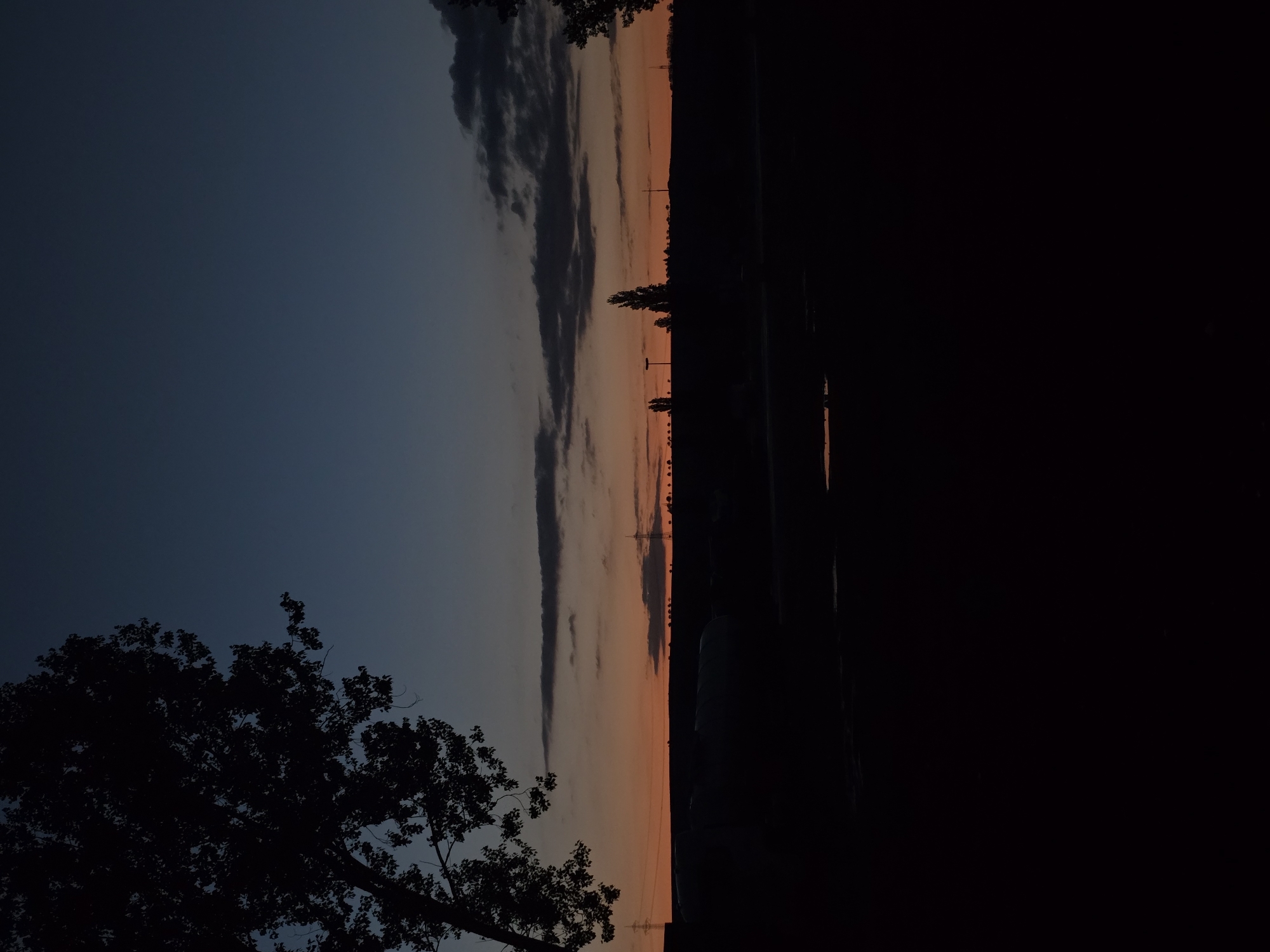 The image depicts a dusk scene in Lutherstadt Eisleben, Germany. The sky is split between a dark blue on the left and orange hues on the right where the sun has set. Clouds streak vertically across the sky. Silhouetted against the sky are trees and power lines. To the right of the image is a large dark area, obscuring details. At the bottom left, a tree branch is visible.