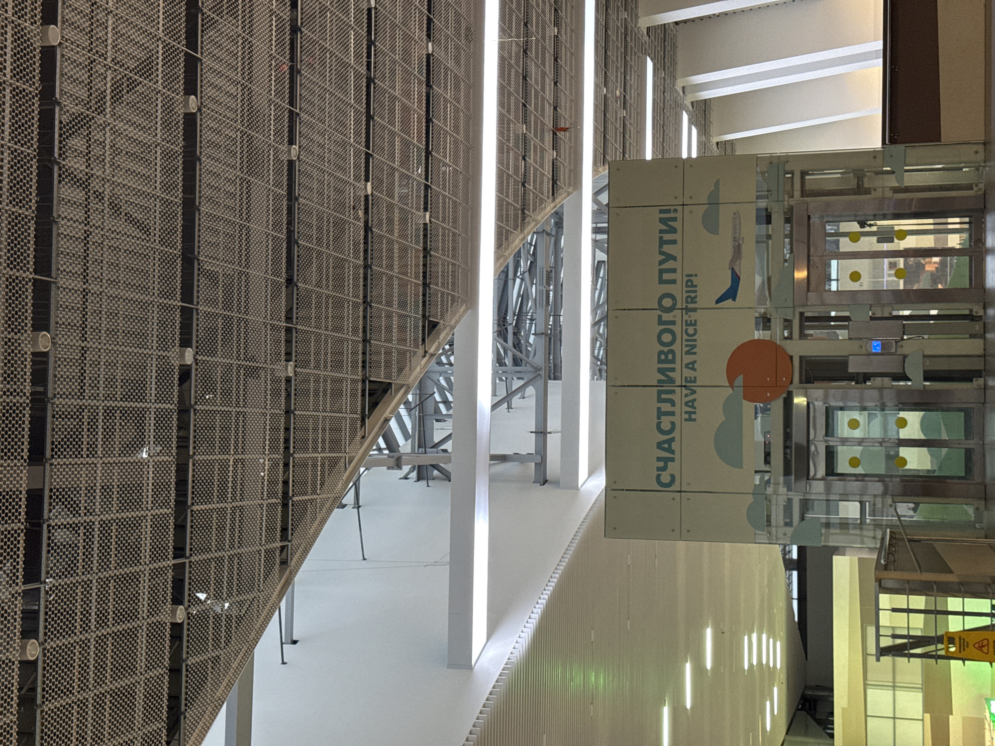 The image depicts an interior space with a perforated metal wall on the left. Parallel to the wall are vertical light columns. A sign with Cyrillic text "СЧАСТЛИВОГО ПУТИ! HAVE A NICE TRIP!" is positioned in the foreground, adjacent to what appears to be a series of elevators or lifts. The setting appears to be an airport or a similar transportation hub. There are no people visible in the frame. The scene is located in Zarechnaya (Gamovskoe S/P), Russia.