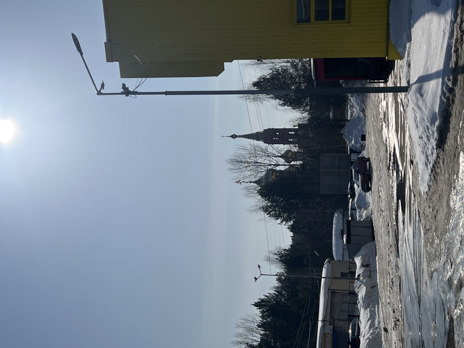 The image displays an outdoor urban environment in Chaikovsky, Russia, under a clear, bright blue sky with a prominent sun flare in the upper right quadrant. The ground in the foreground and midground consists of melting snow, ice, and dark, wet asphalt or dirt, with several puddles reflecting sunlight.
On the right side of the image, a portion of a bright yellow building, likely commercial or industrial, is visible, featuring at least one window. A tall, grey metal streetlight pole stands in front of this building, equipped with a modern luminaire, a smaller secondary light source, and what appears to be a security camera mounted lower on the pole. On the left side, a low-rise, white building with a dark roof and a sign partially displaying "МОНТАЖ" (MONTAGE) is present, with snow accumulated on its roof. Another similar streetlight is visible to its left.
In the midground, a red sedan is positioned on a partially cleared lot, surrounded by melting snow and several dark, utilitarian structures or containers. The vehicle faces towards the left.
The background is dominated by a dense line of evergreen and bare deciduous trees. Rising above the treeline, two distinct structures of the Church of the Holy Great Martyr George the Victorious are visible: a building with two prominent golden onion domes, each topped with an Orthodox cross, and to its right, a reddish-brown brick bell tower with a pointed spire, also crowned with an Orthodox cross. No individuals are present in the scene.