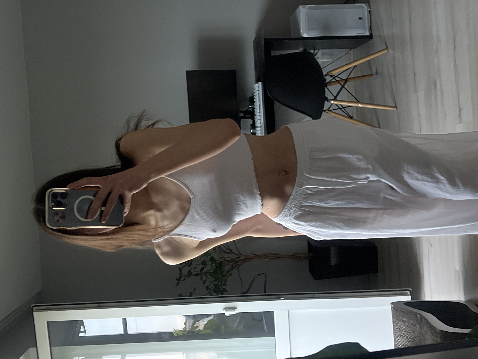 A person stands in an indoor setting, presumably a home, taking a mirror selfie. The person is wearing a white spaghetti-strap top and white pants. A smartphone is held up to obscure their face in the mirror's reflection.
The background contains a window with a view outside. To the right, there is a desk with a computer setup, including a monitor, keyboard, and a black chair. There is a plant visible as well.
The location is not discernible as being in Ternopil, Ukraine.