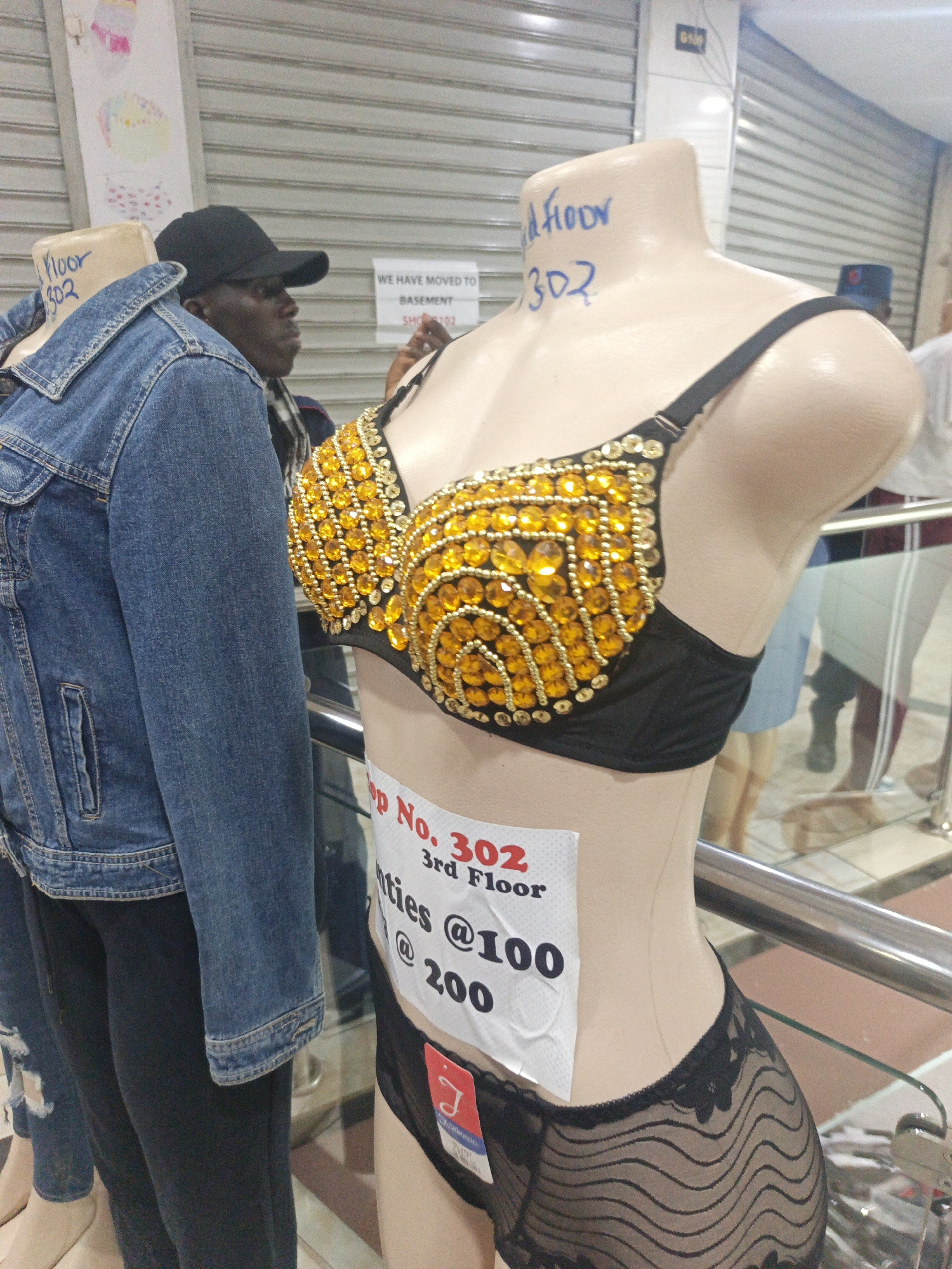 The image shows a mannequin wearing a black bra and panty set embellished with golden rhinestones, displayed inside a shop, likely in Nairobi, Kenya. The mannequin has a sign indicating "Shop No. 302, 3rd Floor" with "Panties @100, @200" written on it. The words "Floor 302" are also written on the mannequin's neck. A man wearing a black cap stands nearby, and a denim jacket is placed on another mannequin to the left. A sign behind the mannequin reads "WE HAVE MOVED TO BASEMENT, SHOP #102." The environment suggests an indoor marketplace or shopping mall.