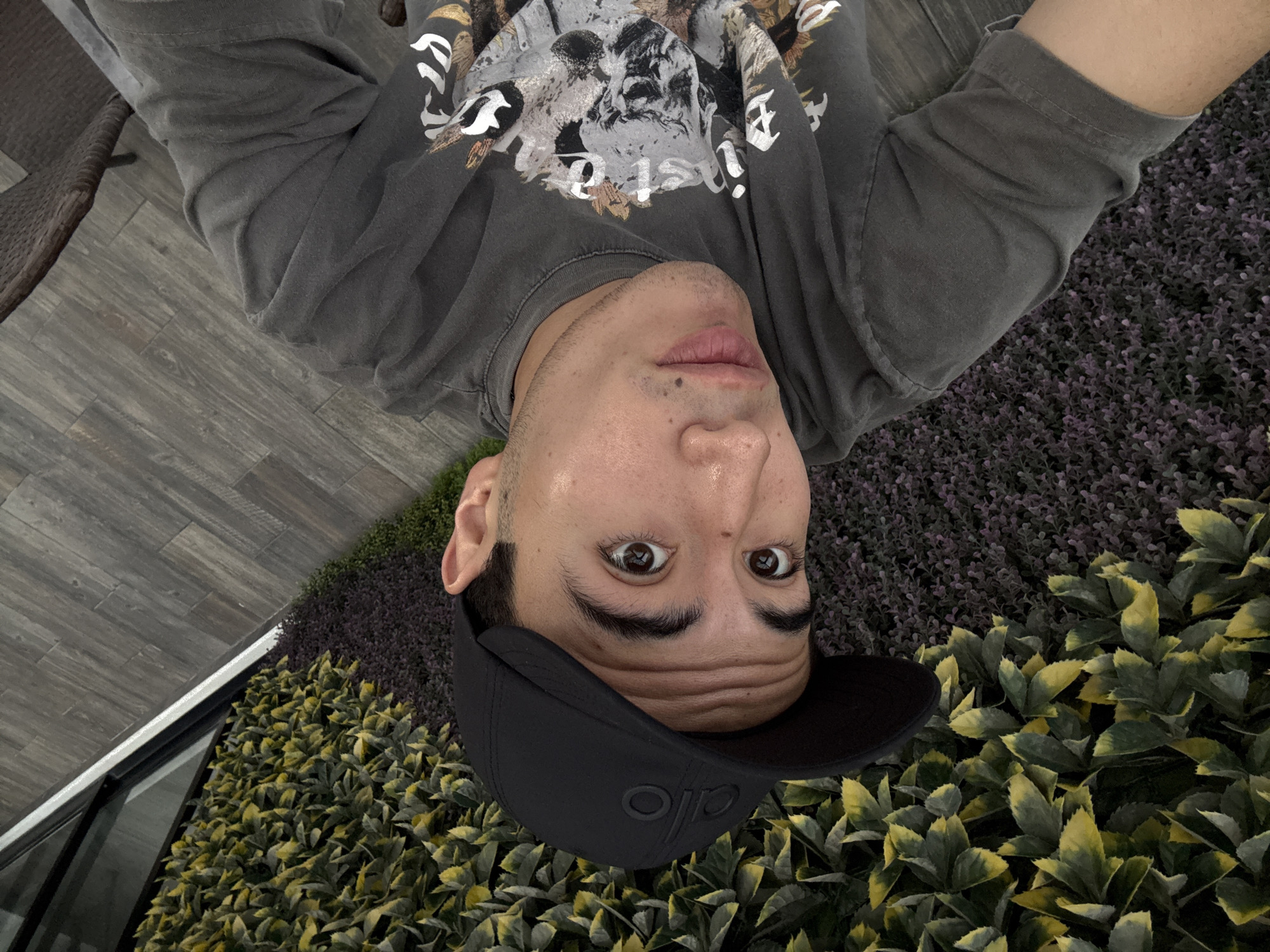 A man wearing a black baseball cap and a grey t-shirt with a skull graphic is taking a selfie outdoors. He is standing in front of a wall of artificial greenery, with a tiled walkway and a wicker chair visible in the background. The scene is set in San Bernardino Tlaxcalancingo, Mexico.