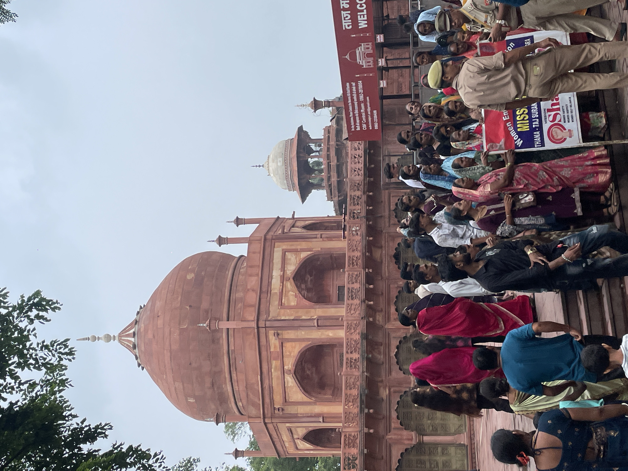 In Agra, India, a group of people, including women in traditional Indian attire such as saris and burqas, stand near a red sandstone monument. Some women hold banners, one of which reads "WOMEN EMPOWERMENT MISSION THANA - TAJ SURAKSHA". Two police officers in uniform are present, one of whom is speaking with a civilian. The environment suggests an outdoor public space, possibly a tourist attraction, with architectural elements typical of Mughal or similar historical structures. The sky is overcast.