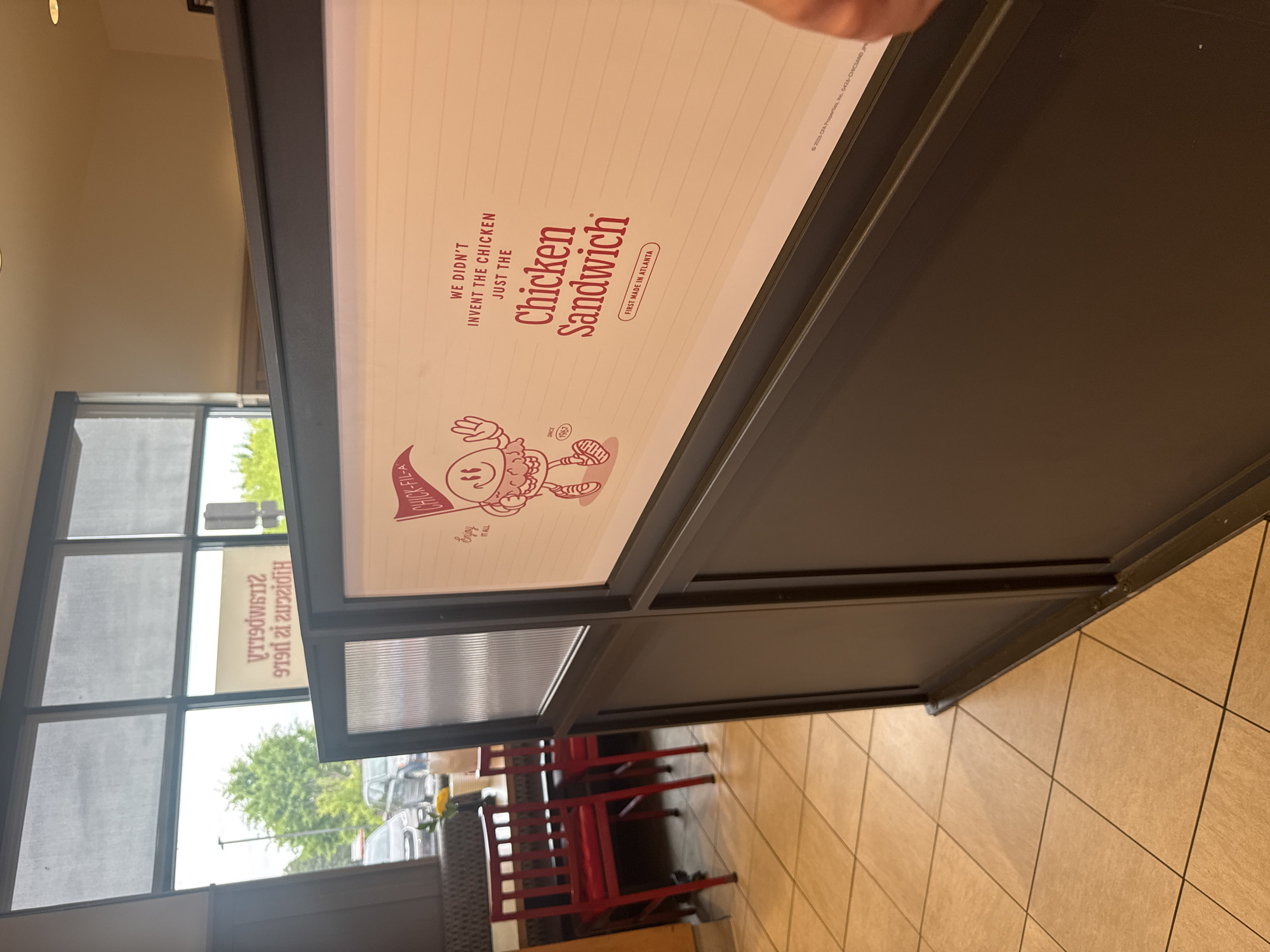 The image shows an indoor scene, possibly within a Chick-fil-A restaurant location in Stone Mountain, Georgia, United States. The foreground features a partition with a prominent advertisement for the Chick-fil-A Chicken Sandwich. The advertisement text reads "We didn't invent the chicken, just the Chicken Sandwich," and "First Made in Atlanta," along with a mascot image. In the background, a window covered in blinds is visible, offering a glimpse of the outside environment, which includes trees and parked cars. Red chairs and tables can be seen through the partition, indicating a seating area. The floor appears to be tiled.