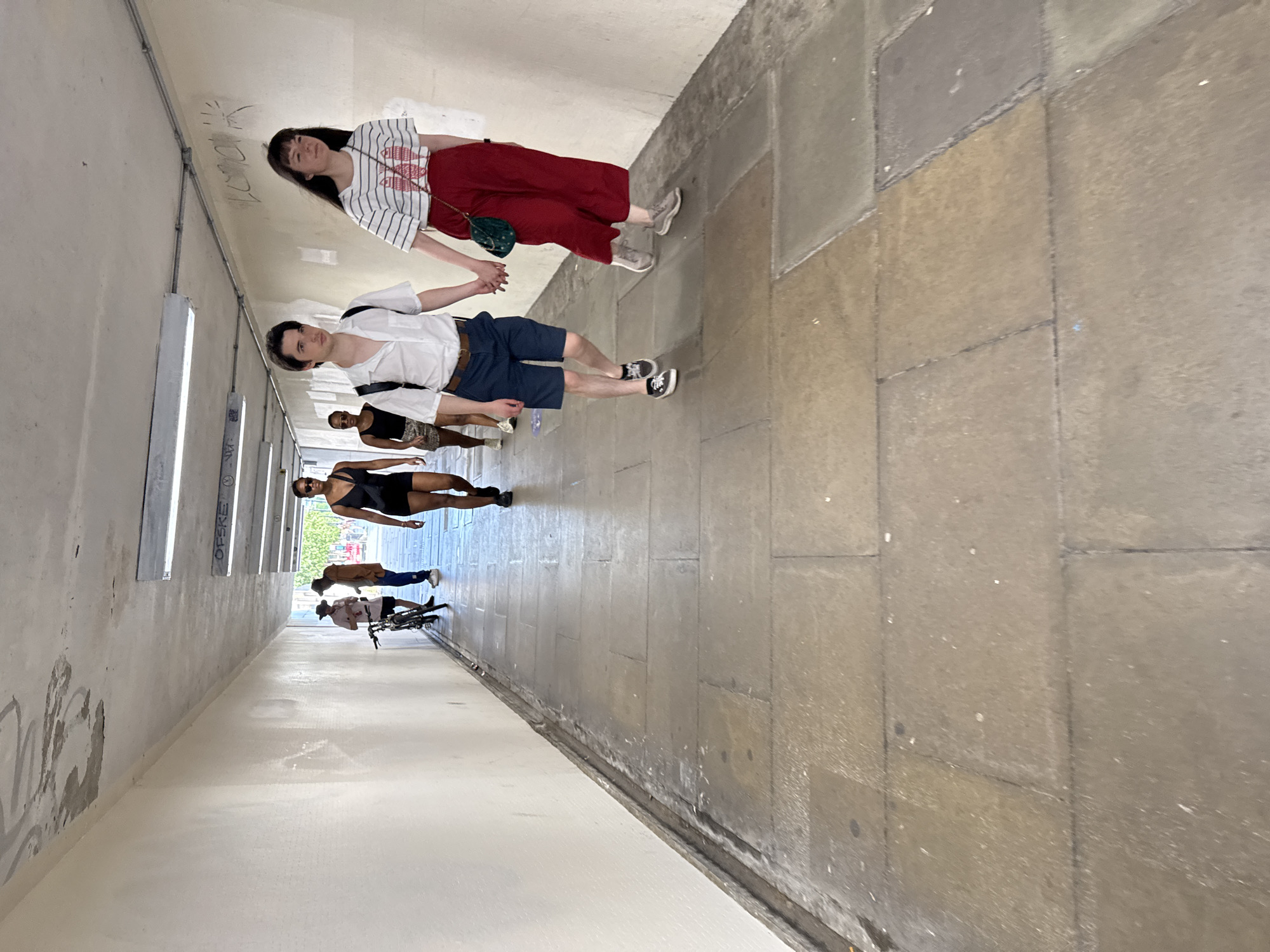 The image depicts a street underpass with a white, textured ceiling and walls, illuminated by fluorescent lights. The ground is paved with large, rectangular stone tiles. Several individuals are walking through the underpass. In the foreground, a young couple walks hand-in-hand. The male is wearing a white short-sleeved shirt, navy shorts, and dark shoes. The female is wearing a striped t-shirt and red wide-leg pants, carrying a small green bag.
Further back, a person in a black swimsuit and sunglasses walks away from the camera. Behind them, another person in a black tank top and patterned skirt is visible. To the left, a group of three individuals stands near a bicycle. One person in a pink shirt and a hat is facing them, and another person in a brown jacket and dark pants is also present. The underpass appears to be in a public area, possibly a pedestrian walkway or thoroughfare within the city.
Based on the architectural style, the paving stones, and the general atmosphere, this underpass is highly likely to be in London, United Kingdom.