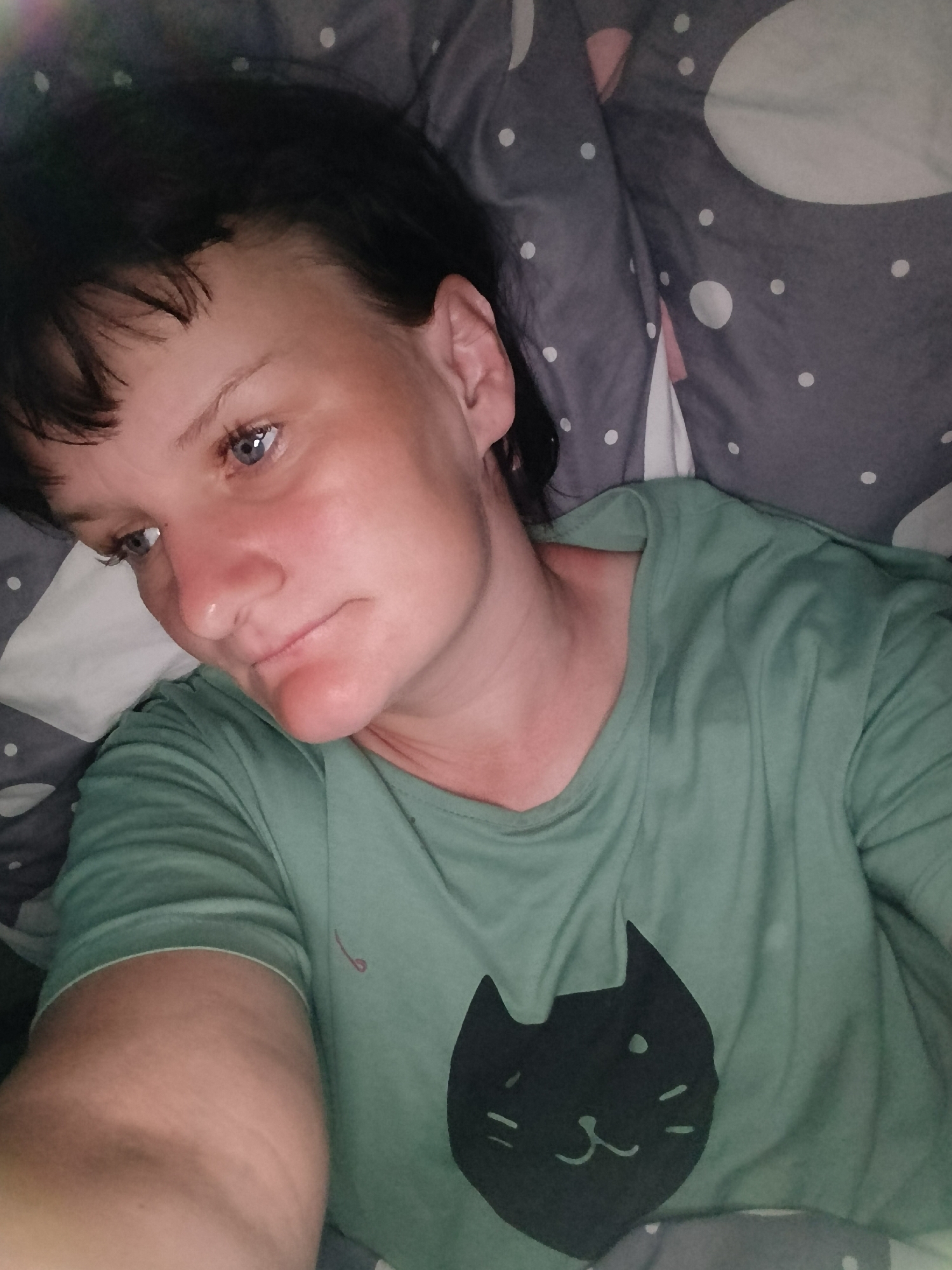 A woman with short, dark hair and blue eyes is lying in bed. She is wearing a light green t-shirt with a black cat face design. The bed linens are grey with white polka dots and white and pink moon shapes. The lighting is dim. Location: Фоки, Russia (implied by user request).