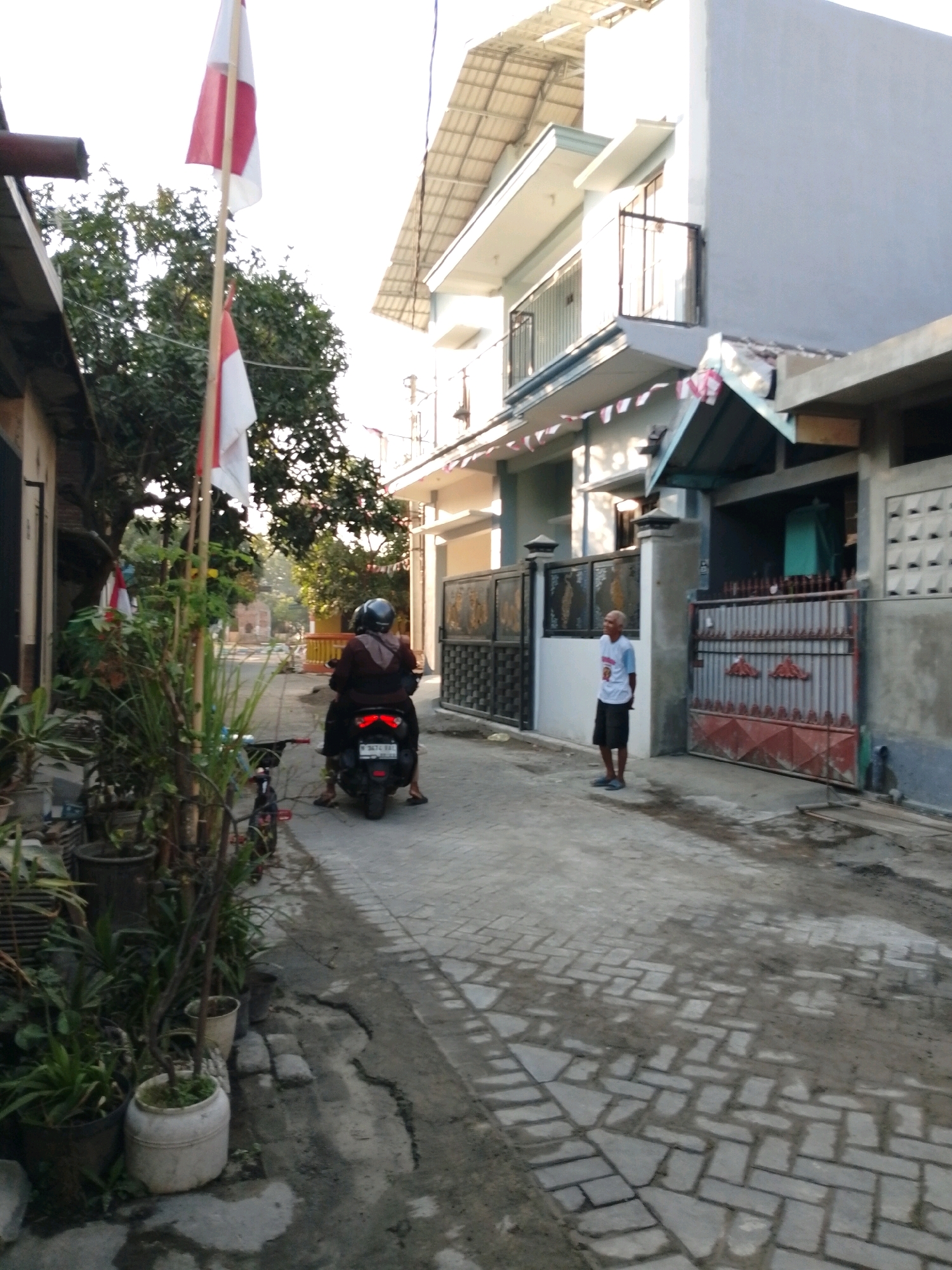 In a narrow, paved alleyway in Pasuruan, Indonesia, a person wearing a helmet rides a scooter, their back to the camera. The scooter has a license plate visible. To the left, a tall flag pole flies the Indonesian flag, accompanied by a smaller flag and various potted plants. On the right side of the alley, a light blue, two-story building stands with a balcony and barred windows. A man in a white t-shirt and dark shorts stands near a large metal gate in front of the building. The alley is lined with fences and appears to be a residential area. The time of day is likely morning or late afternoon, given the soft lighting.